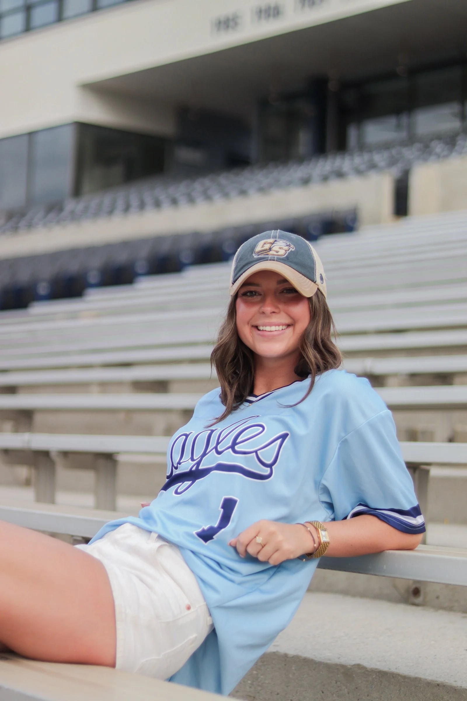 Smiling woman sitting on stadium bleachers wearing a baseball cap and a light blue sports jersey.