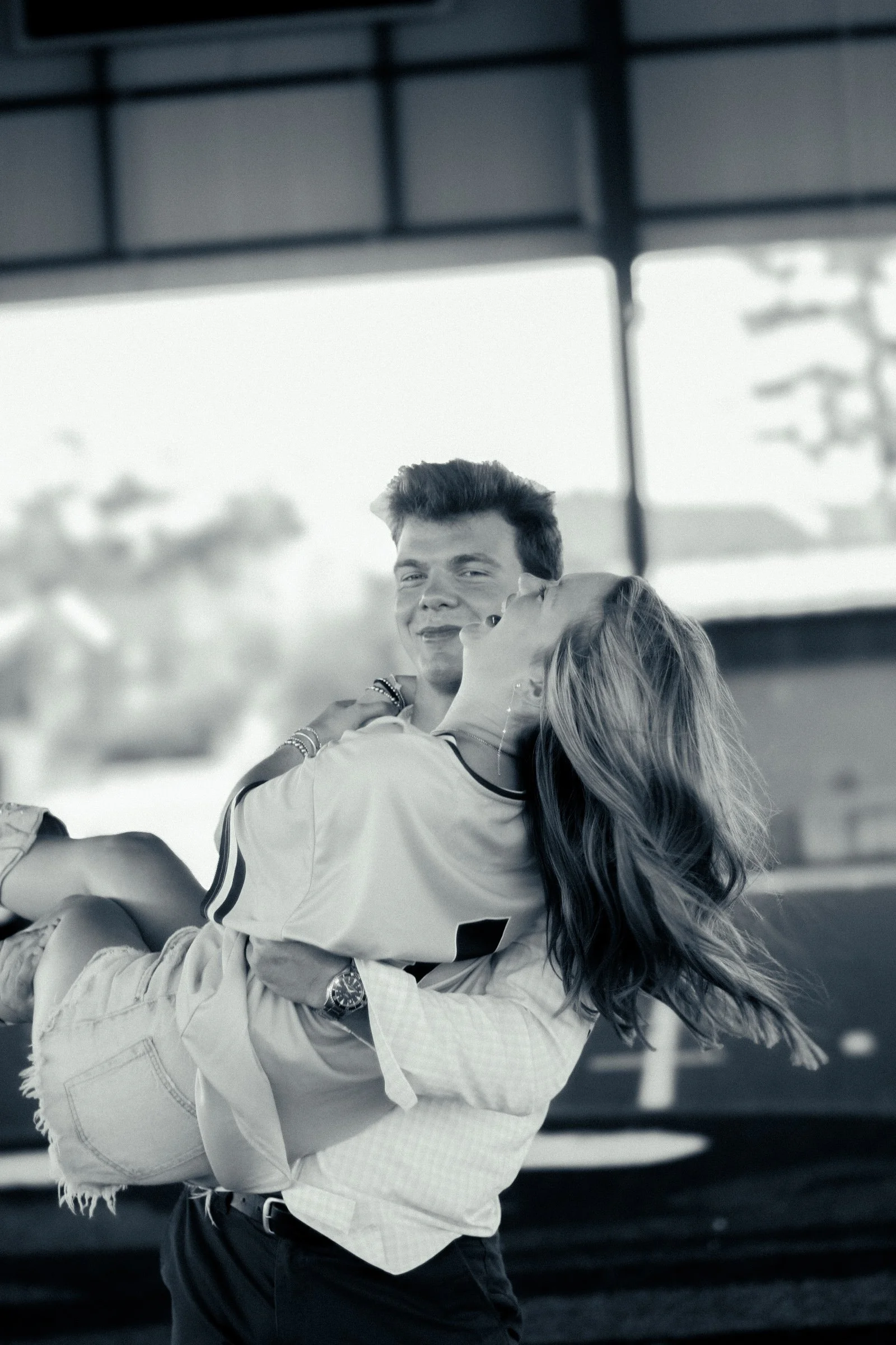 A young man dancing with a young woman in an outdoor setting, with the man lifting the woman in his arms, both smiling. The background includes a large structure and cloudy sky.