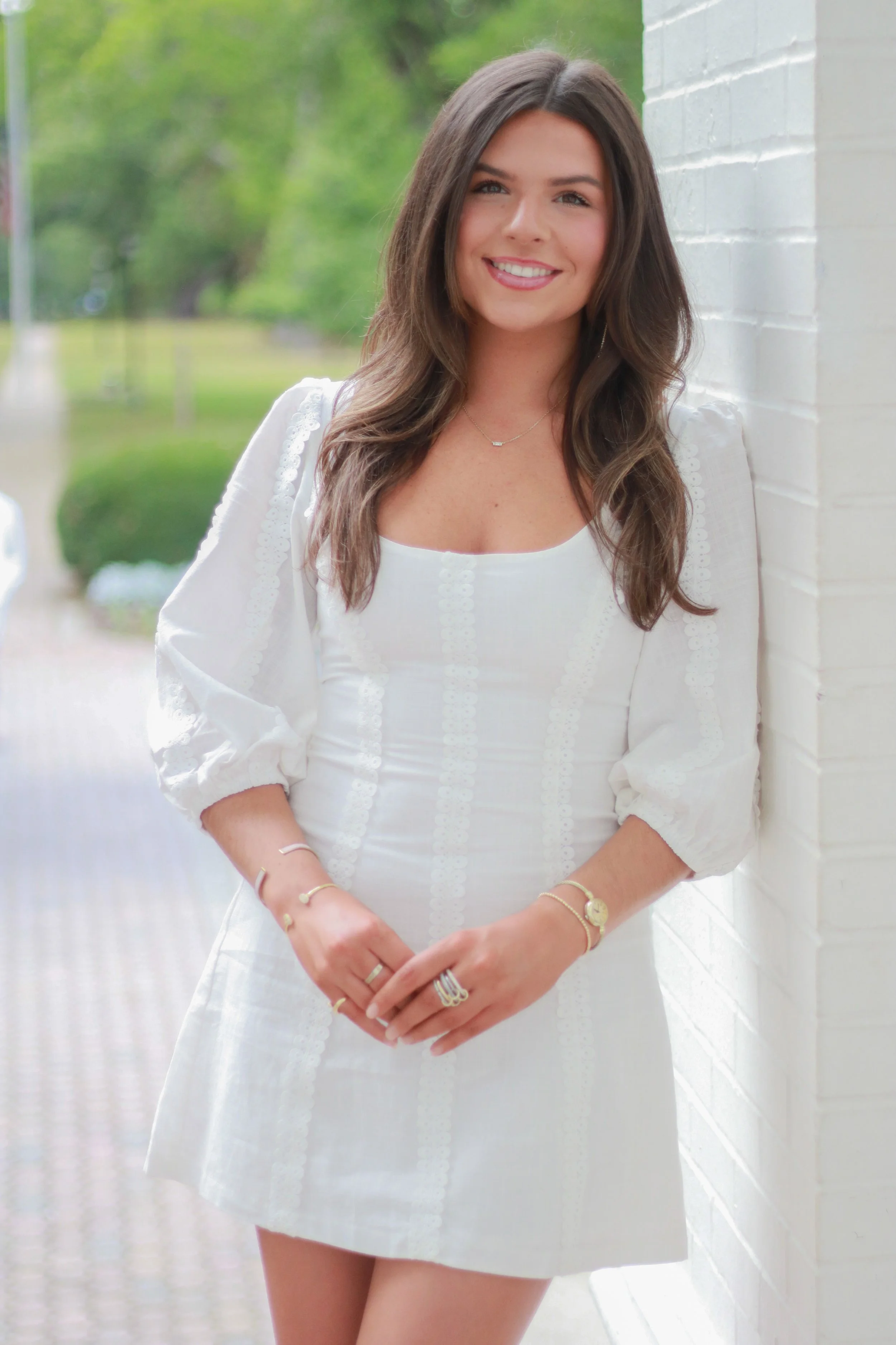 Young woman with long brown hair, wearing a white dress and gold jewelry, standing outdoors at a white brick wall, smiling.