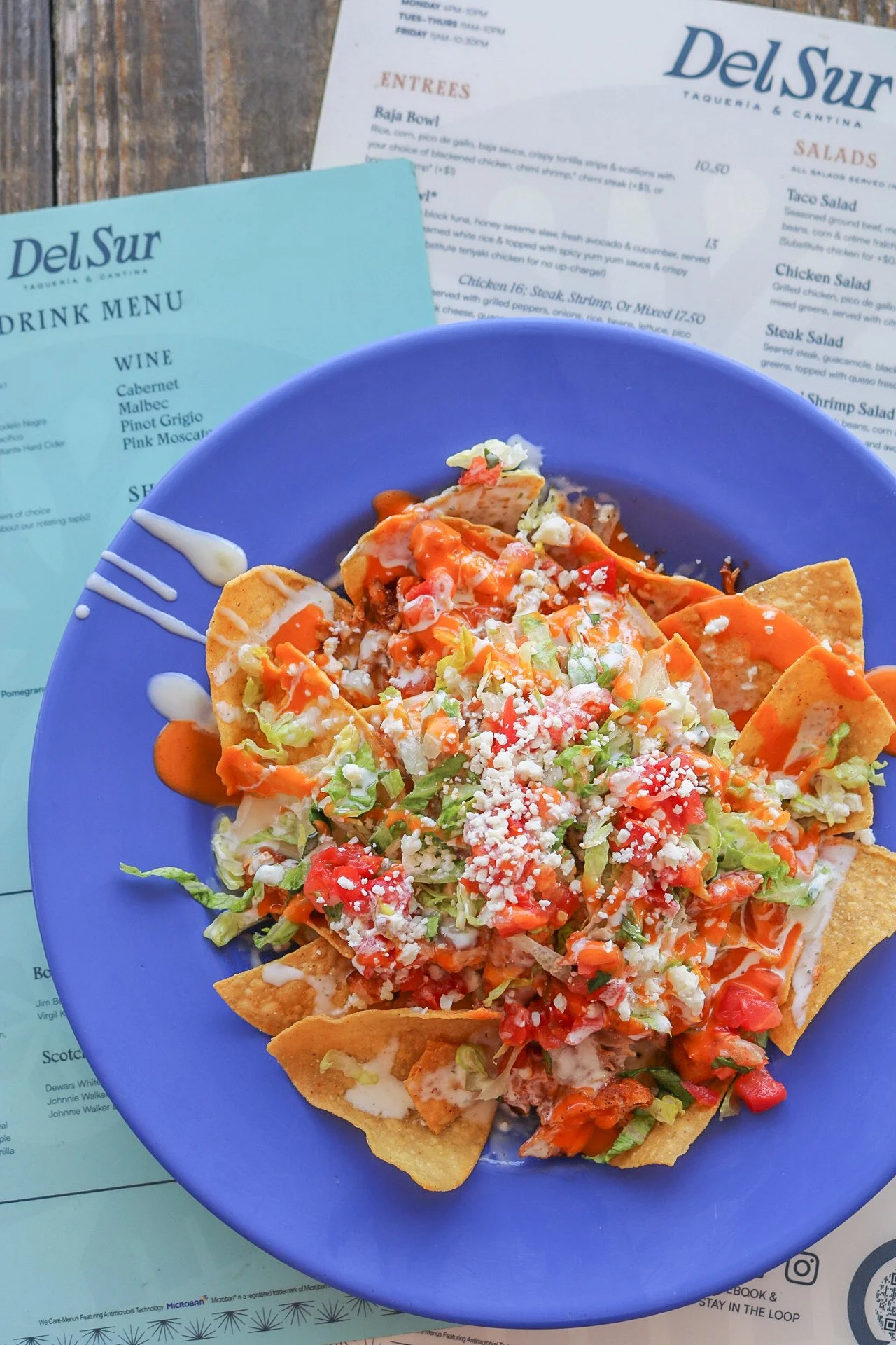 A plate of nachos topped with shredded cheese, chopped lettuce, diced tomatoes, sour cream, and hot sauce on a blue plate, with restaurant menus in the background.