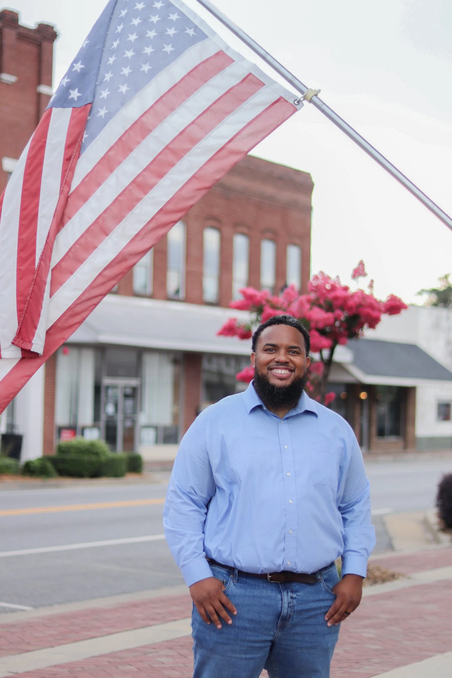 A man standing outdoors in front of a brick building and pink flowering tree, wearing a blue dress shirt and jeans, smiling at the camera with an American flag on a pole behind him.