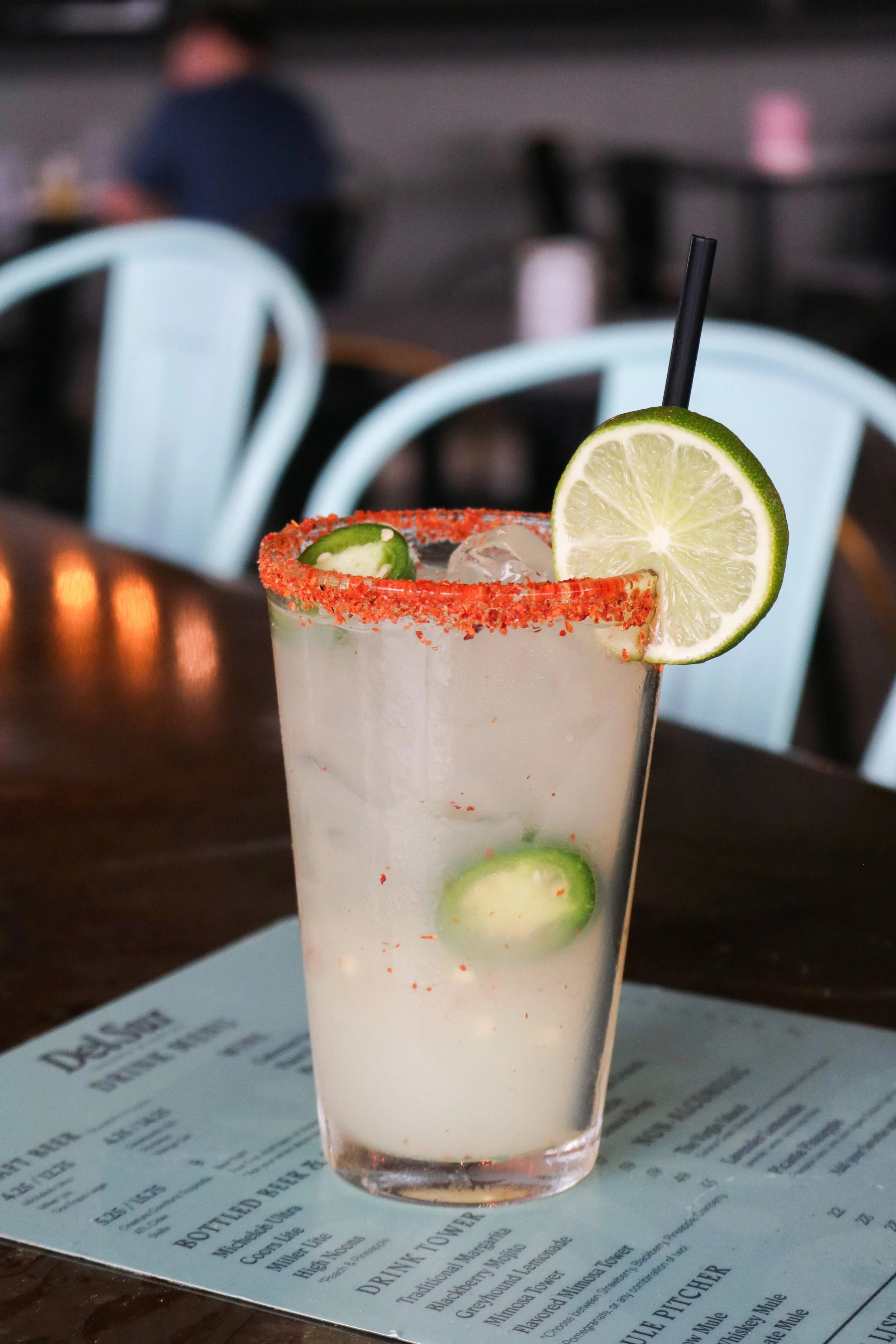 A drink in a tall glass garnished with a lime slice and rim dipped in chili powder, with lime slices inside, on top of a menu on a table in a restaurant or bar.