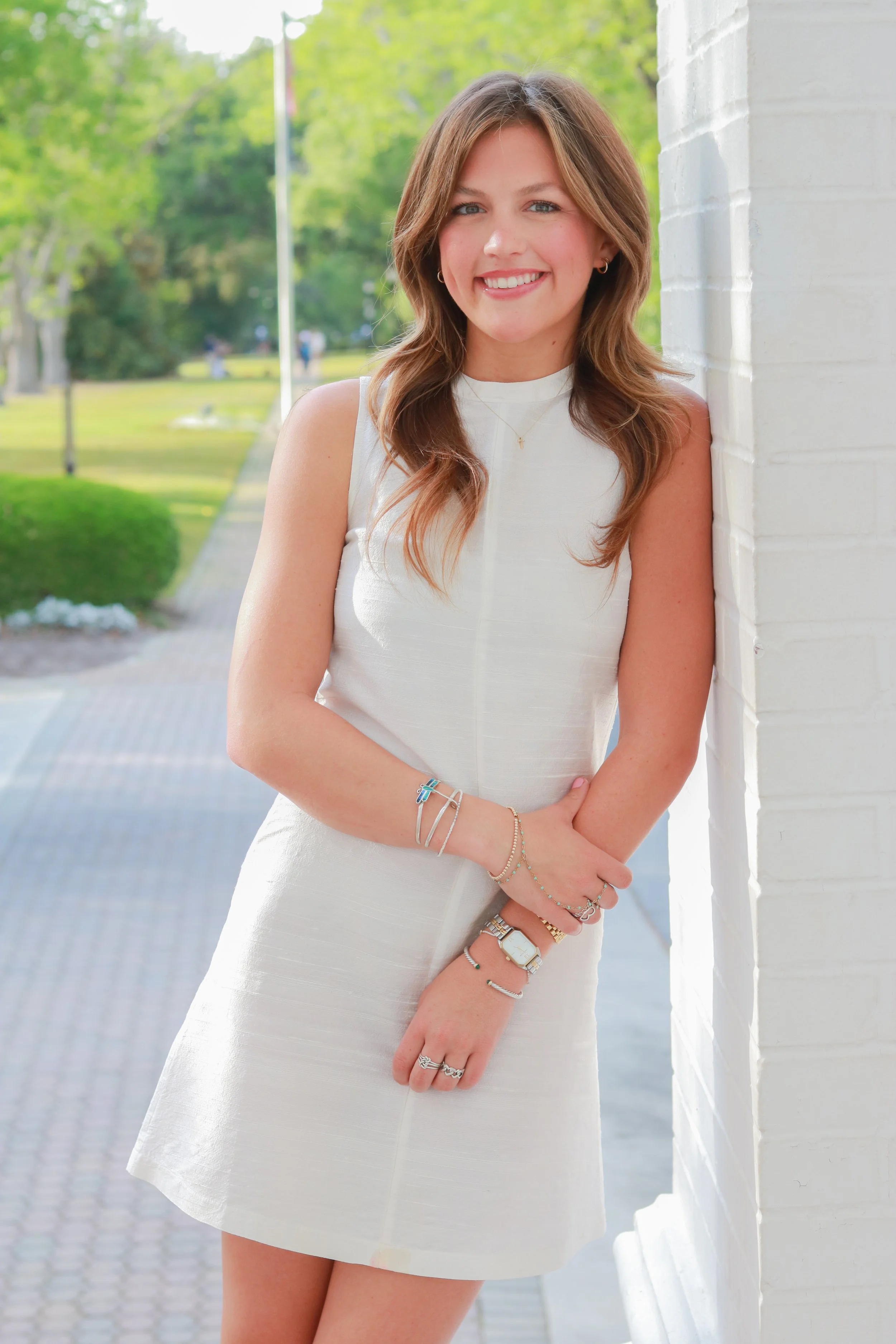 A young woman with light brown hair smiling, wearing a sleeveless white dress and jewelry, standing outdoors near a white brick wall with greenery and trees in the background.