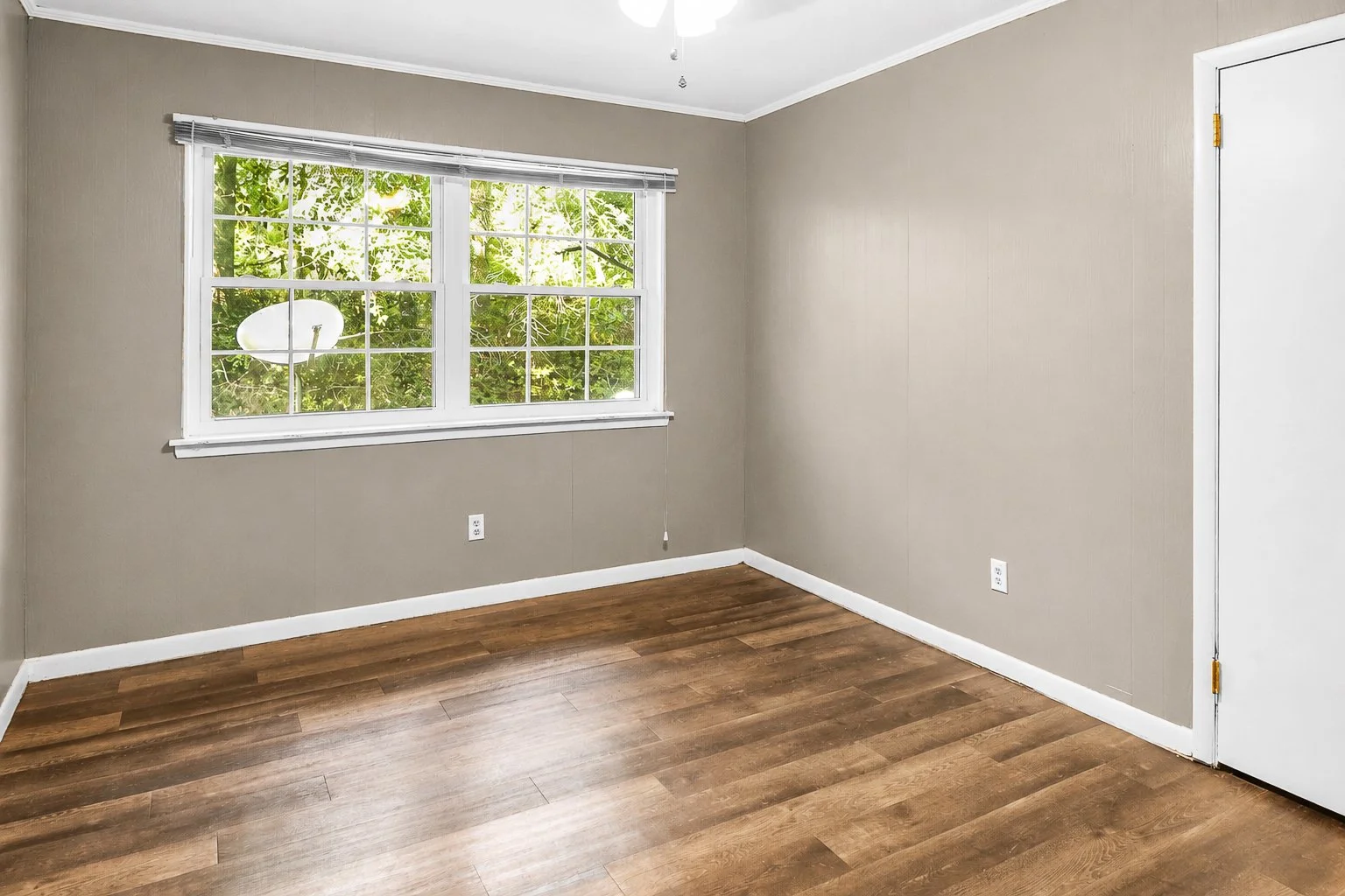 Empty room with beige walls, hardwood floor, white window with blinds, and a white door.