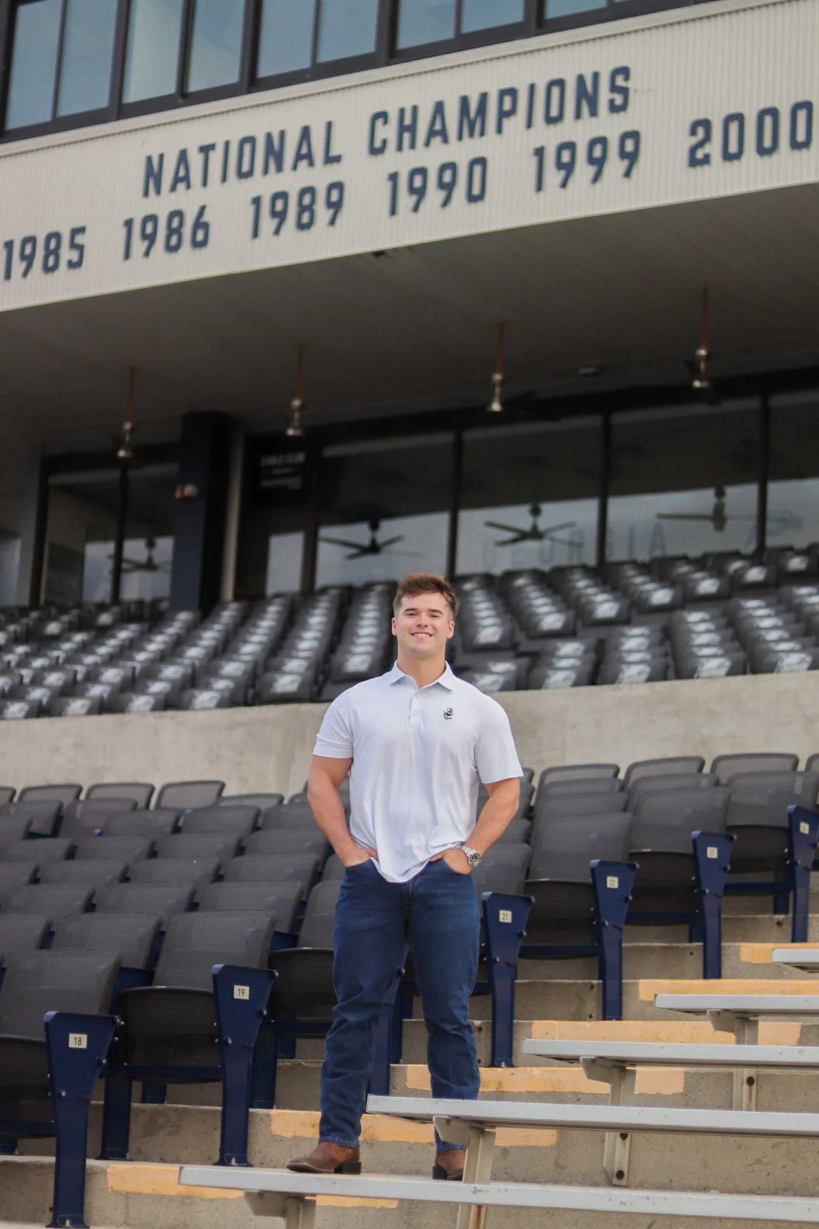 A man wearing a light-colored shirt and dark jeans standing on stadium seats with a banner above him listing years of national championships.