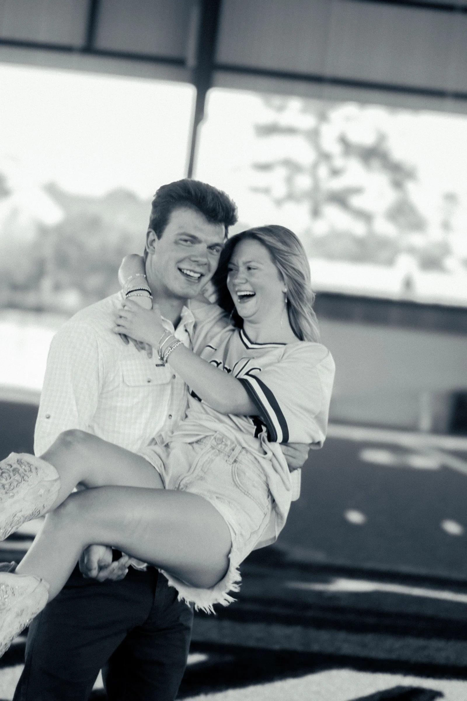 A young man lifting a young woman in his arms, outdoors with a cloudy sky and trees in the background, both laughing and looking happy.