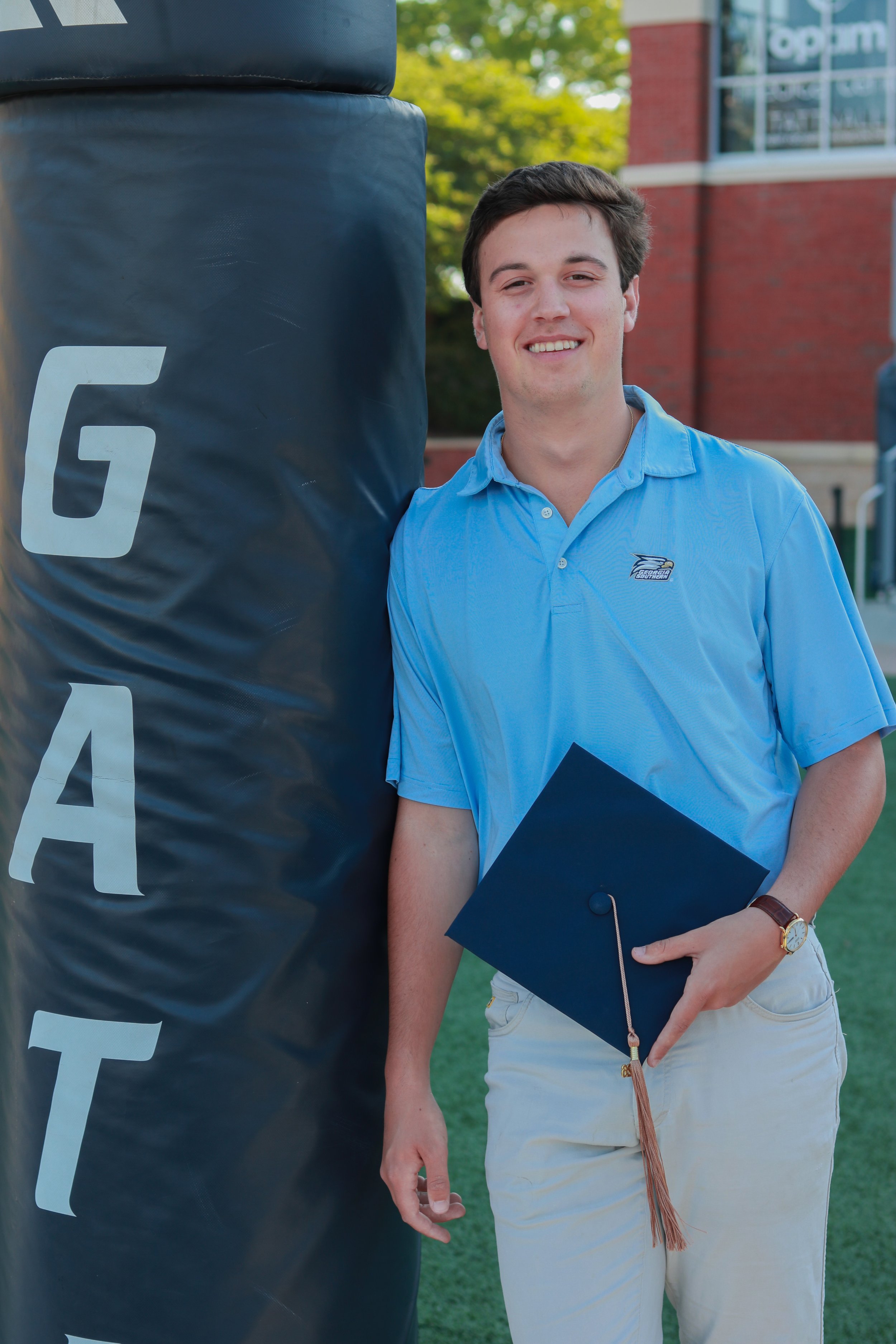Smiling young man in a blue polo shirt holding a graduation cap, standing outdoors beside a large black volleyball with white lettering, in front of a brick building with windows.