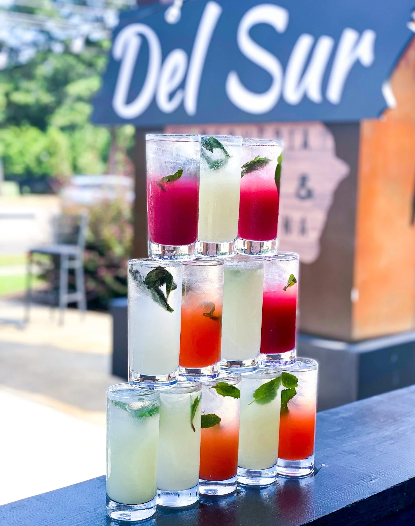 Six tall cocktail glasses filled with colorful drinks, garnished with mint leaves, arranged on a black surface outdoors with a blurred background and a sign reading 'Del Sur'.