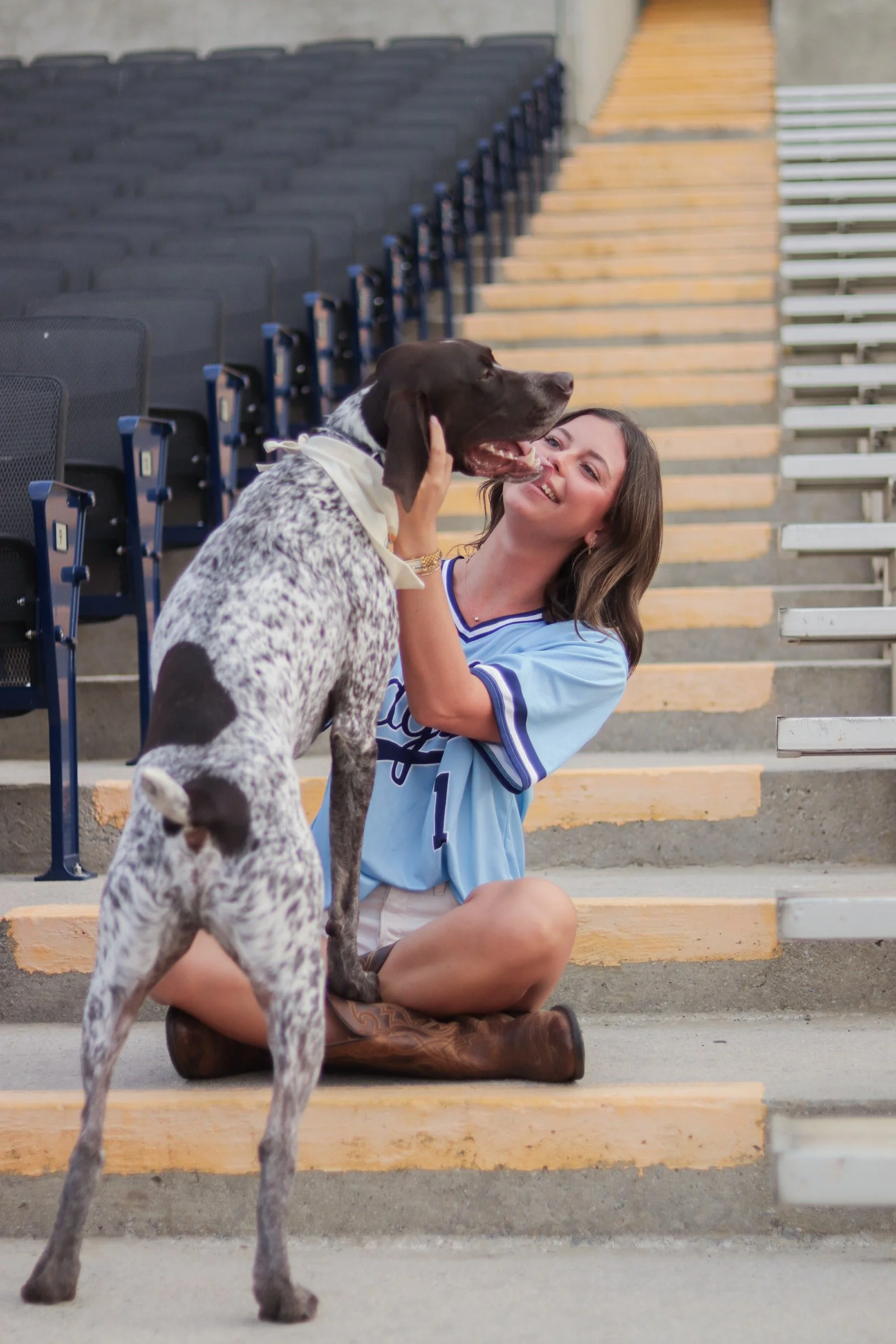 A young woman sitting on steps smiling and holding a large eyes brown and white German Shorthaired Pointer dog, which is standing on its hind legs with its tongue out. Empty chairs are visible in the background.