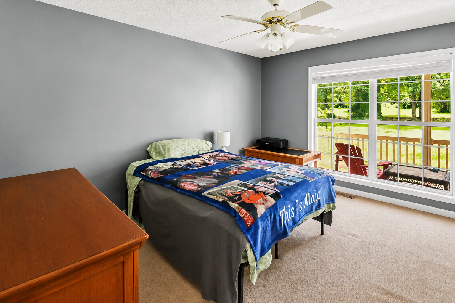 A bedroom with gray walls and a large window showing a view of green trees and a porch with red chairs. There is a bed with a blanket featuring people and the words "This Is Maia," a small white lamp, a wooden dresser, and a black printer on a wooden