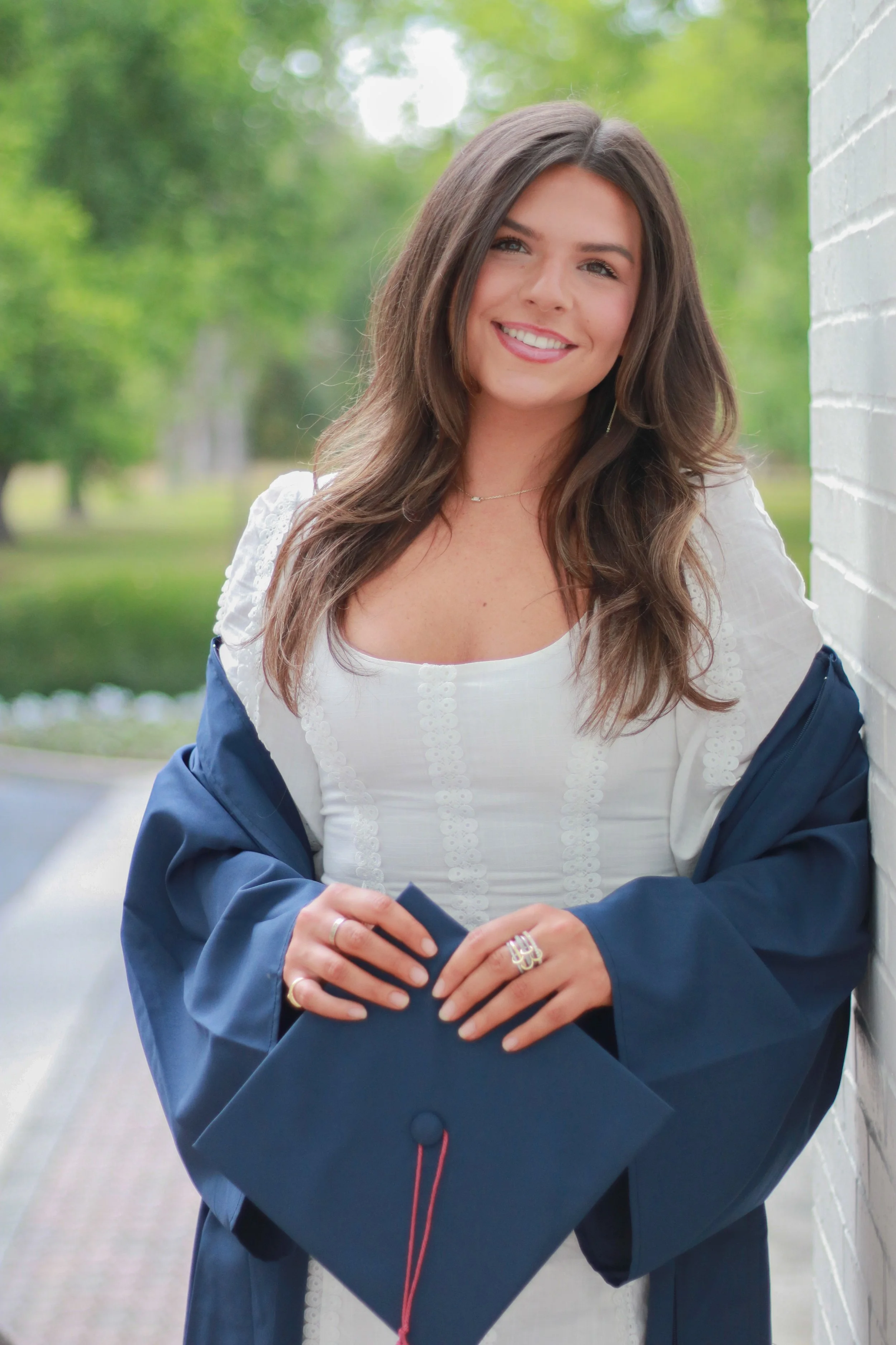 Young woman with long brown hair smiling, wearing a white top and a blue jacket, holding a navy blue folder, standing outdoors near a white brick wall with a blurred green park background.