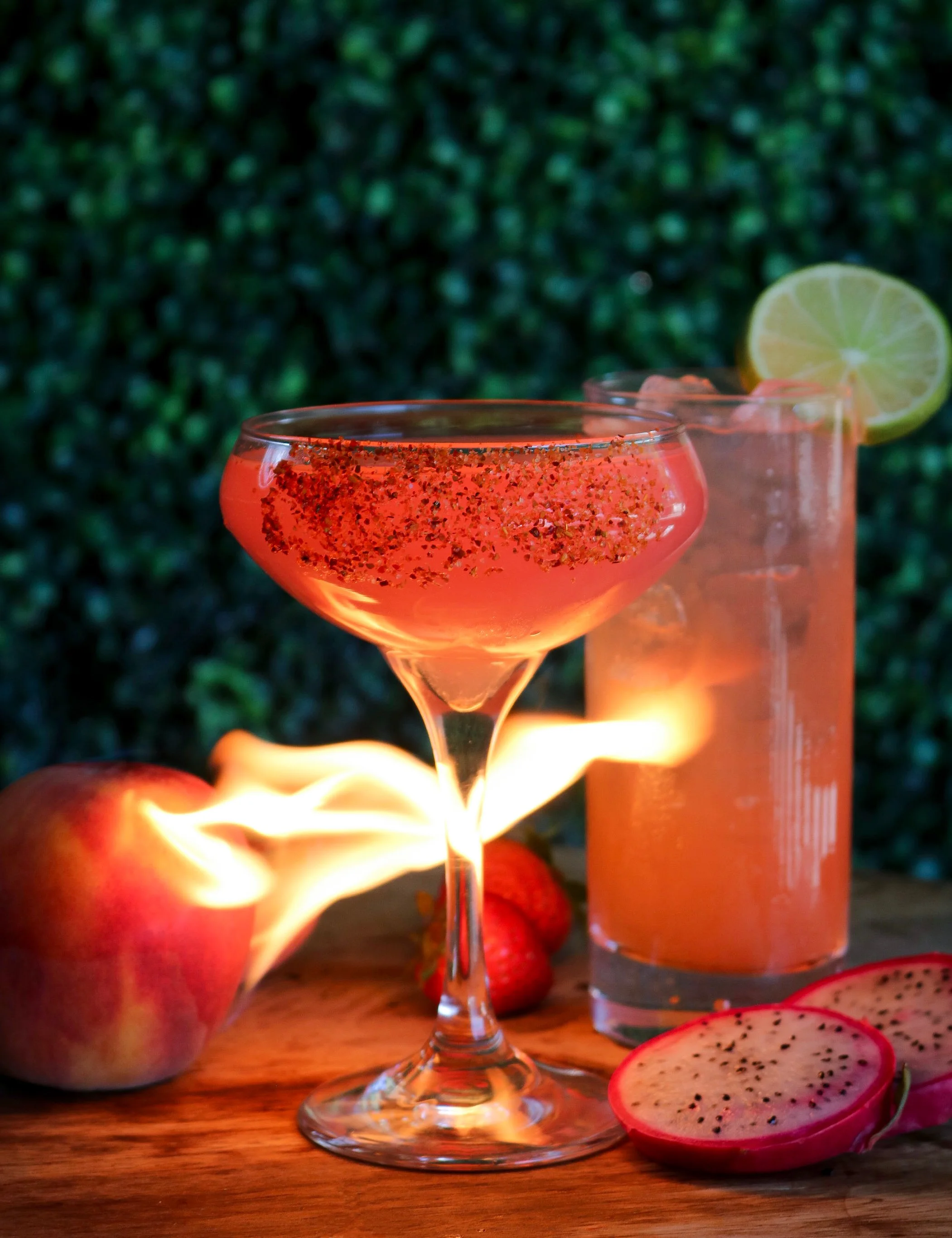 A pink cocktail in a stemmed glass with chili flakes on rim, sliced lime garnish, and a tall pink drink behind. Red apples and dragon fruit slices are on a wooden surface with greenery background.