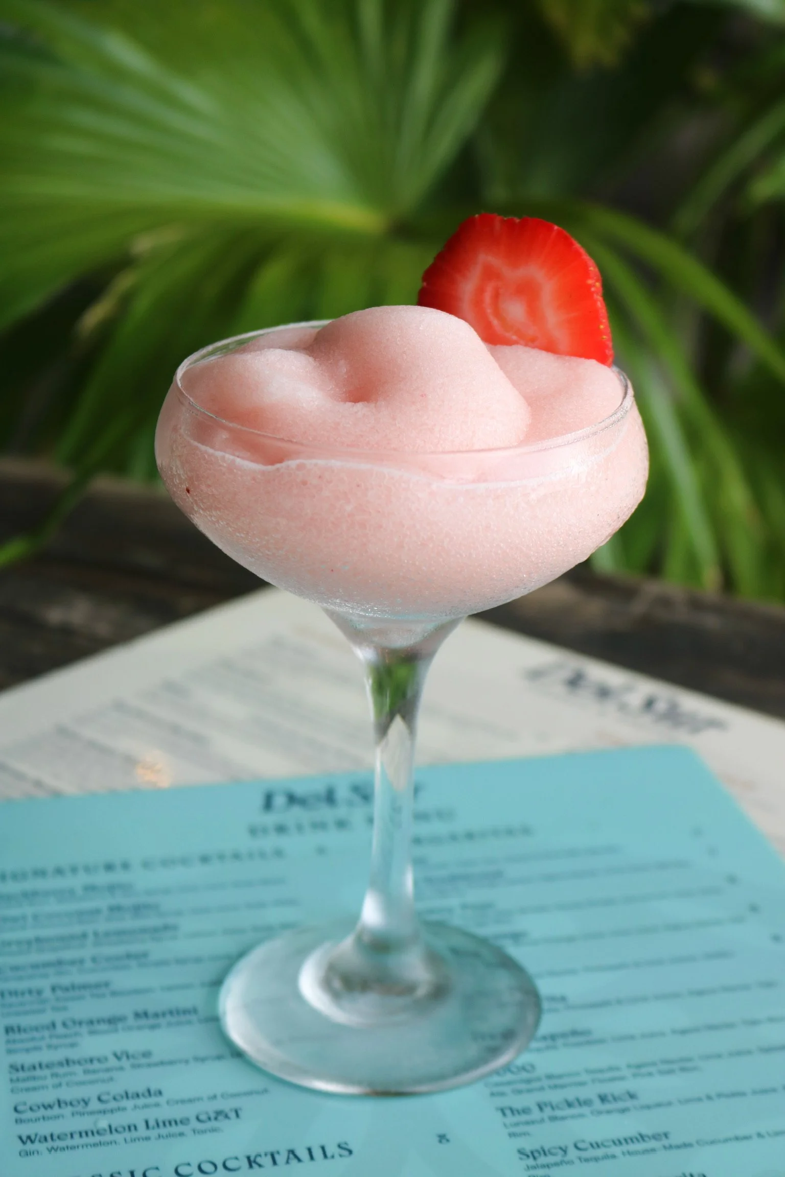 A pink sorbet in a glass with a slice of strawberry on top, placed on a menu in an outdoor setting.