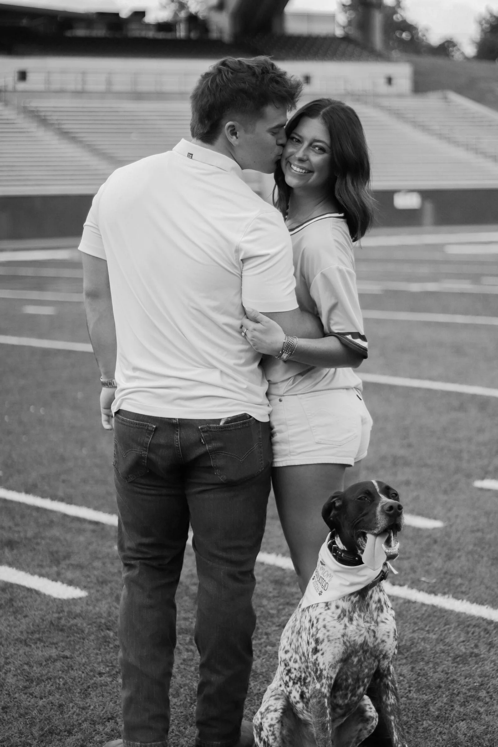 A black and white photo of a young man and woman embracing on a sports field, with a dog sitting nearby. The young man is kissing the woman's cheek while she smiles at the camera.