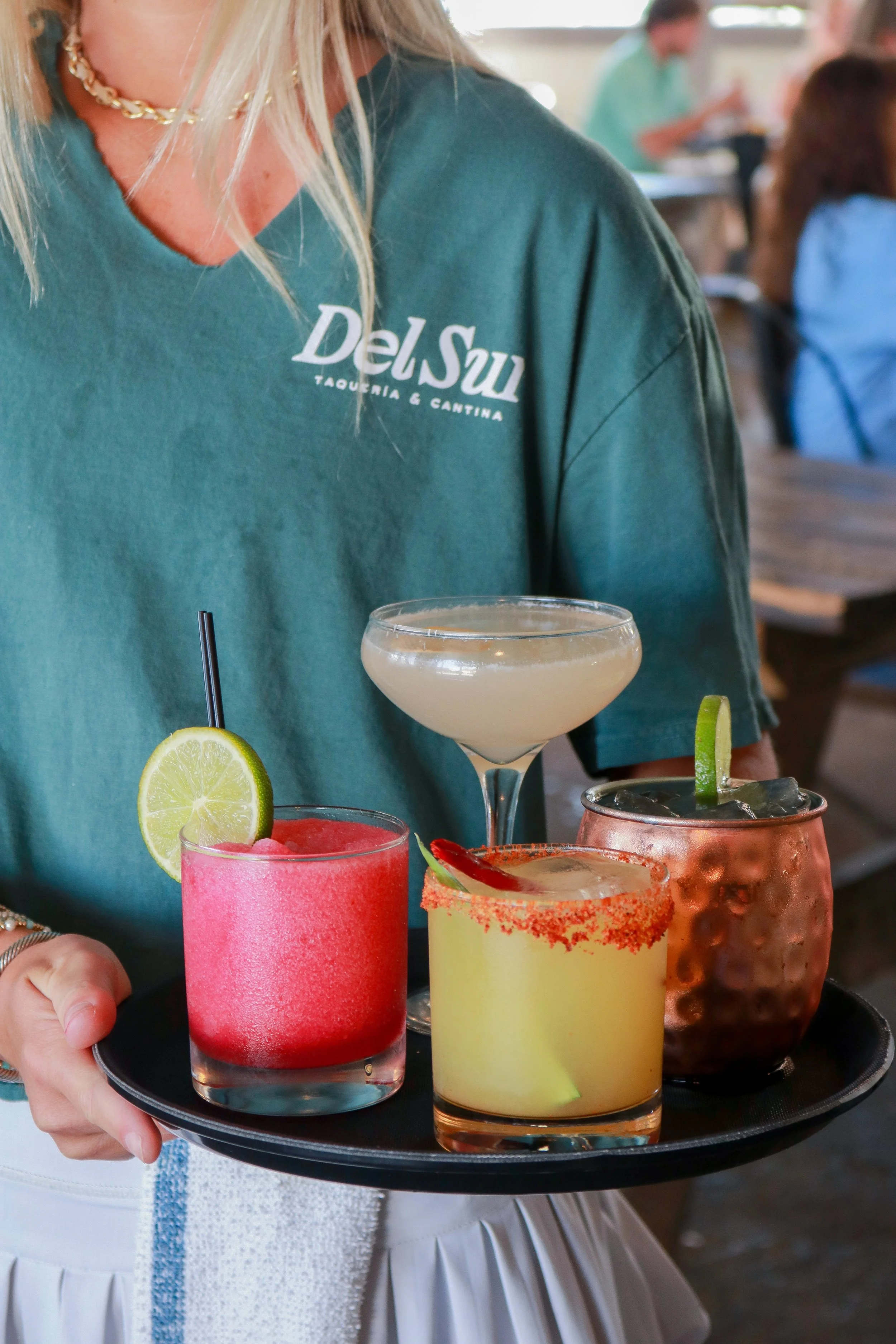A woman holding a black tray with various colorful cocktails, including a pink drink with a lime wedge, a yellow drink with a salted rim, a white cocktail, and a copper copper mug, at a bar or restaurant.