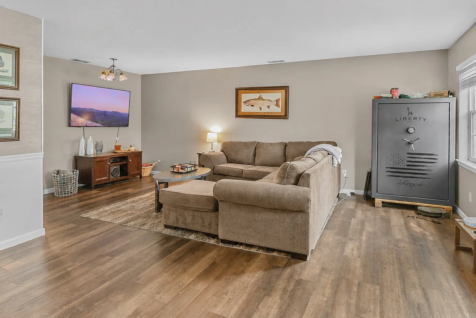 Living room with beige sectional sofa, wall art, wooden flooring, a TV mounted on the wall, a wooden cabinet below the TV, a small coffee table, a floor lamp, and a large gray safe.