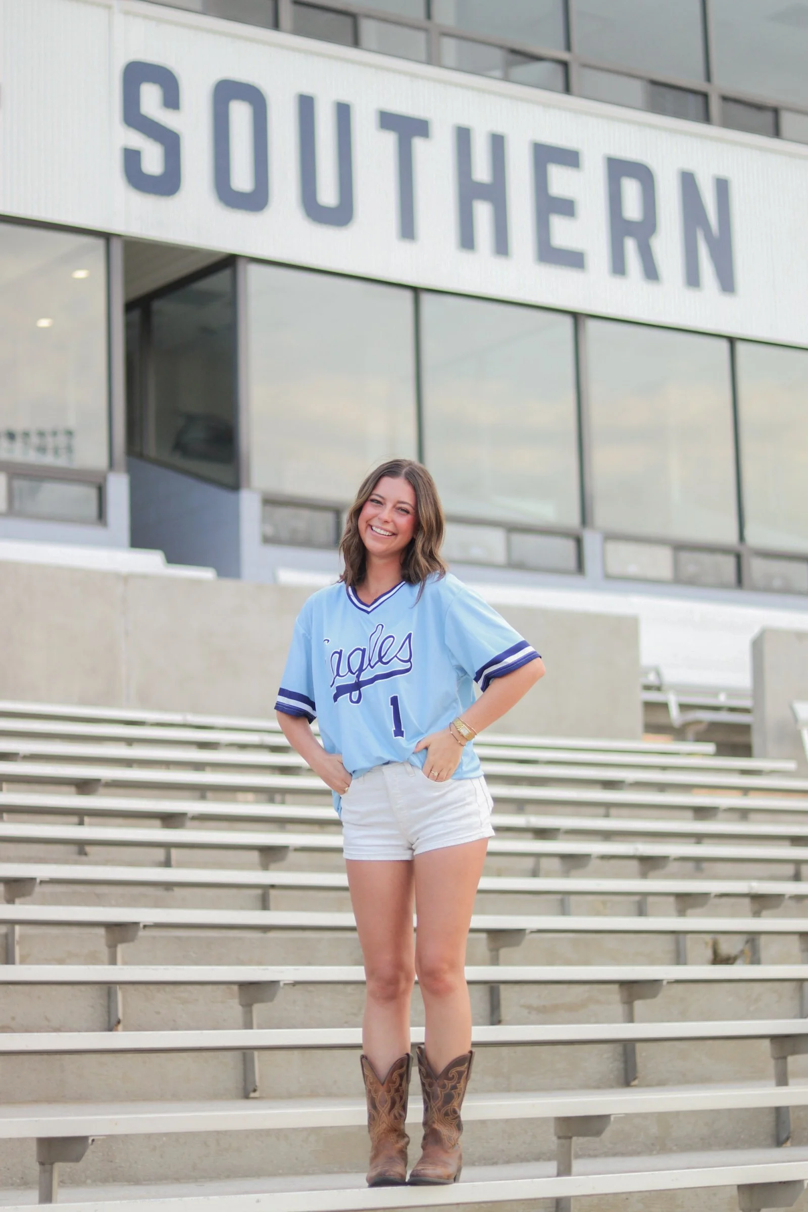 A woman standing on stadium bleachers in front of a modern building with a sign that reads 'Southern,' wearing a light blue sports jersey, white shorts, and cowboy boots, smiling at the camera.
