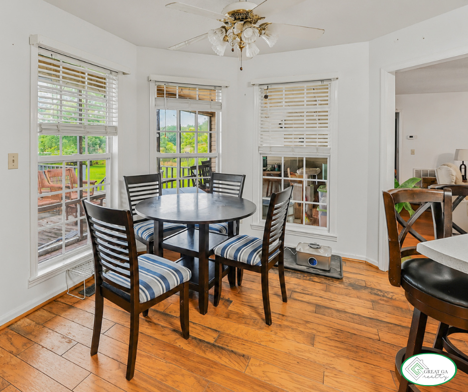 Dining area with a round black table, five wooden chairs with striped blue and white cushions, hardwood flooring, and windows showing a green outdoor landscape. Ceiling fan with lights and a small pet exercise device on the floor.