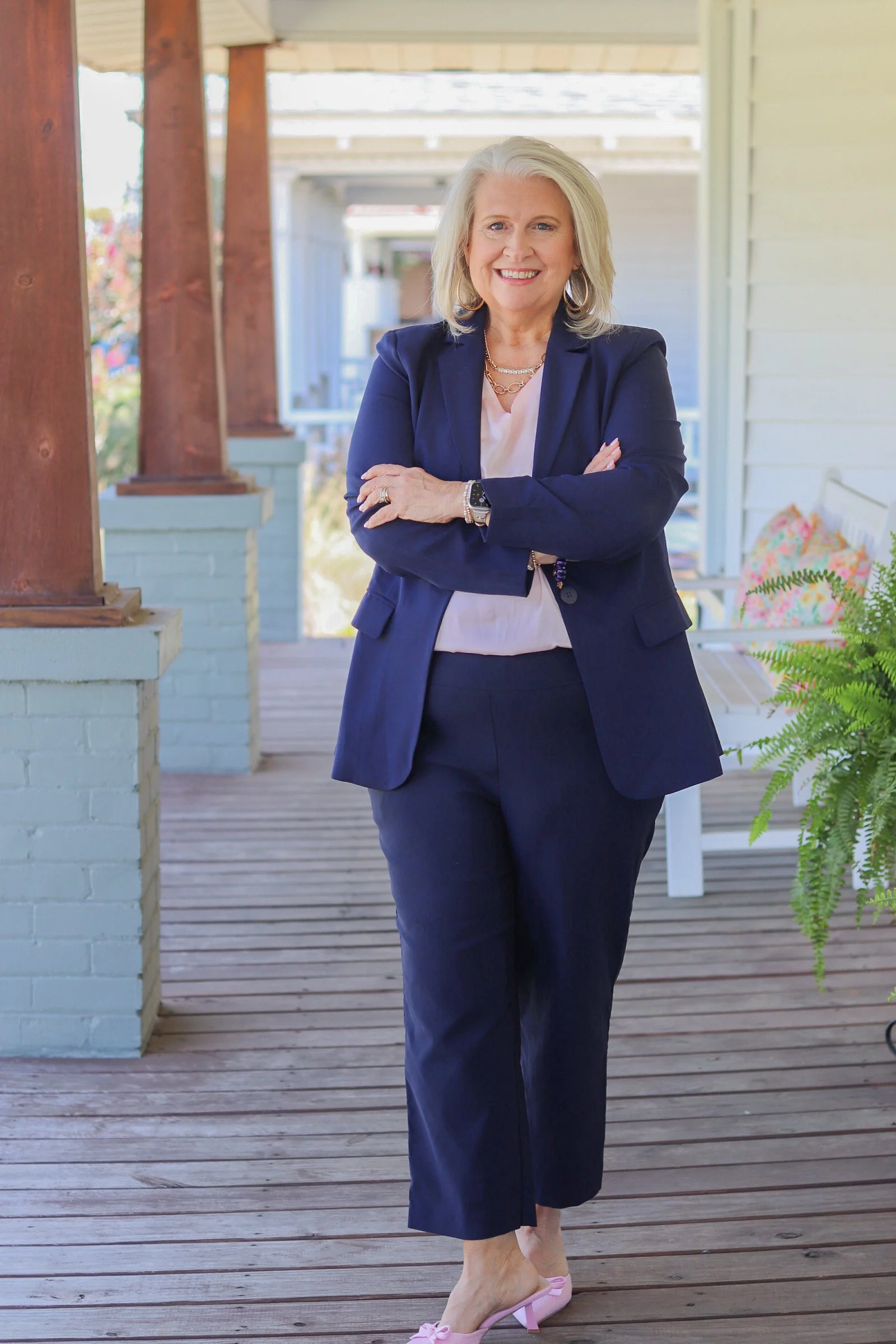 A woman standing on a wooden porch, smiling, dressed in a navy blazer and pants, wearing jewelry, and pink heels.