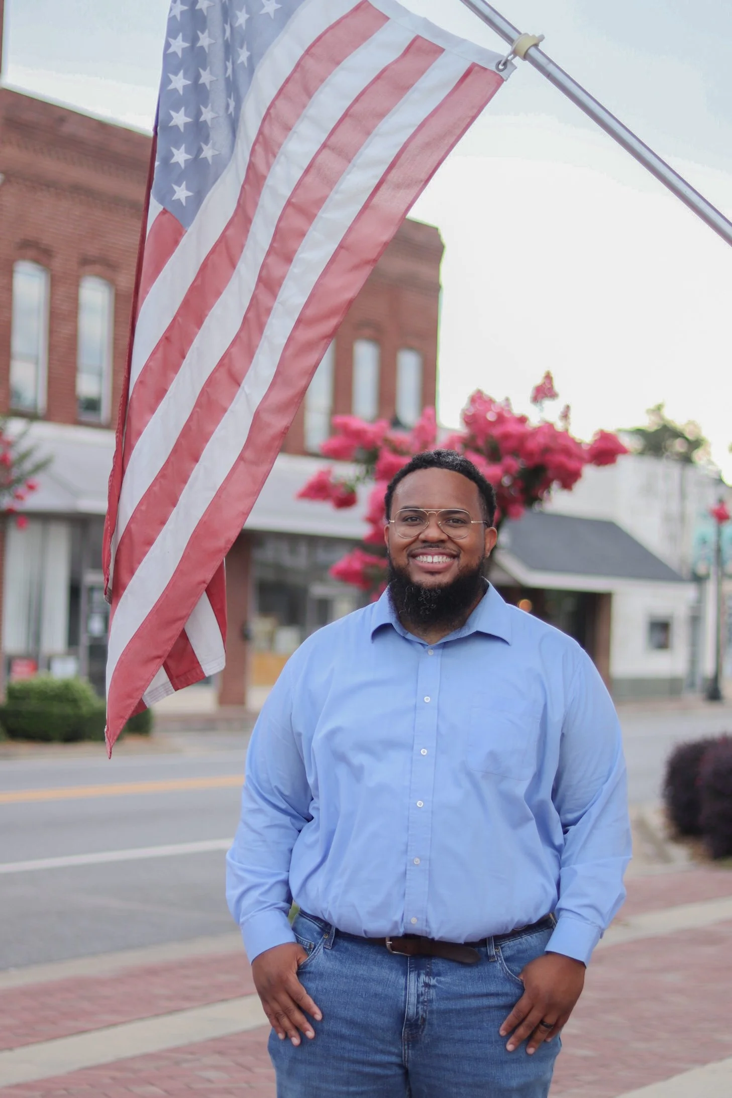 A man standing outdoors during daytime, smiling, next to an American flag on a pole. The background includes a brick building, pink flowers, and a small tree.