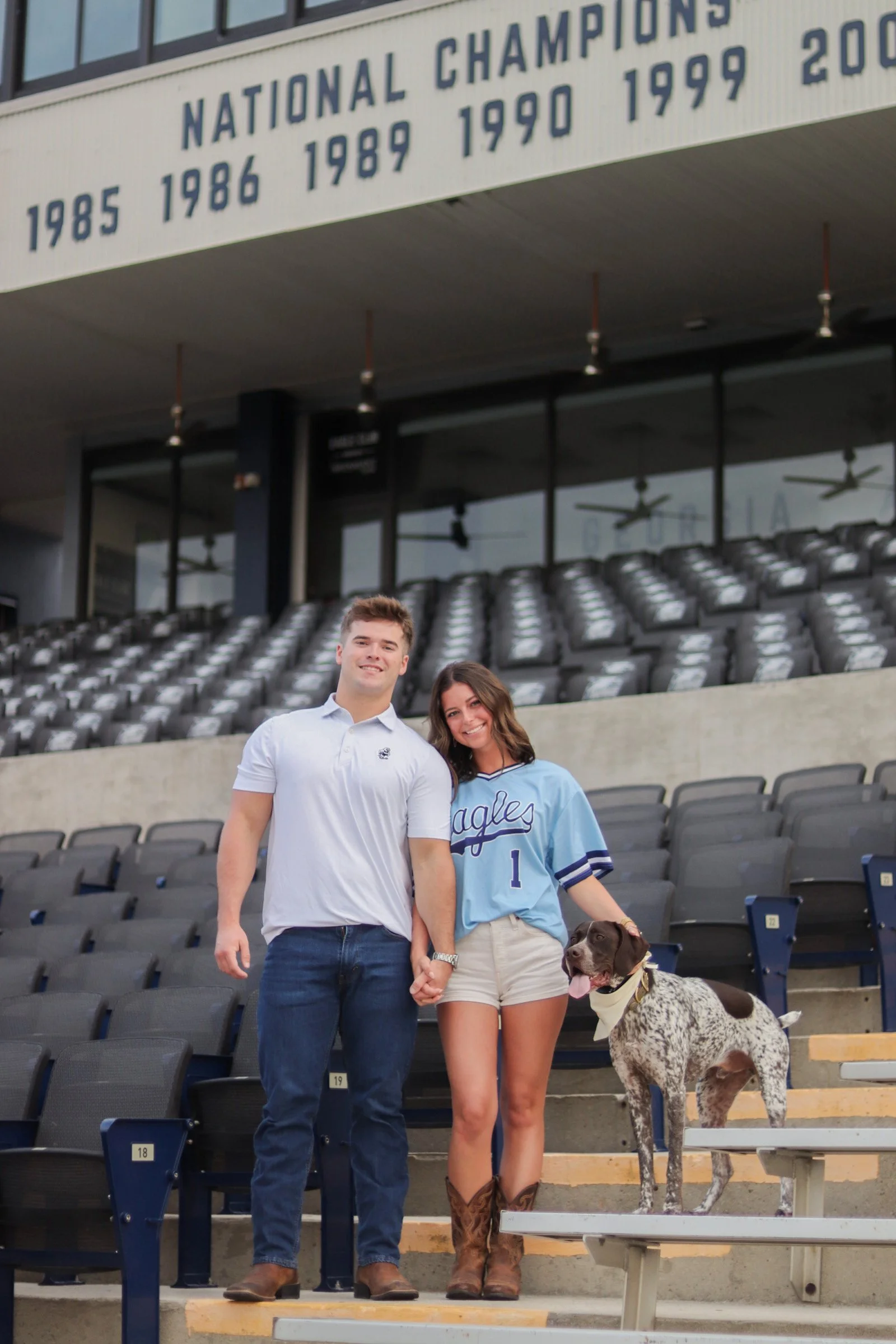 Two young adults, a man and a woman, standing on stadium steps with their dog, a large dalmatian, at a sports stadium. The woman is wearing a light blue sports jersey, and the man is wearing a white shirts. They are holding hands and smiling at the c