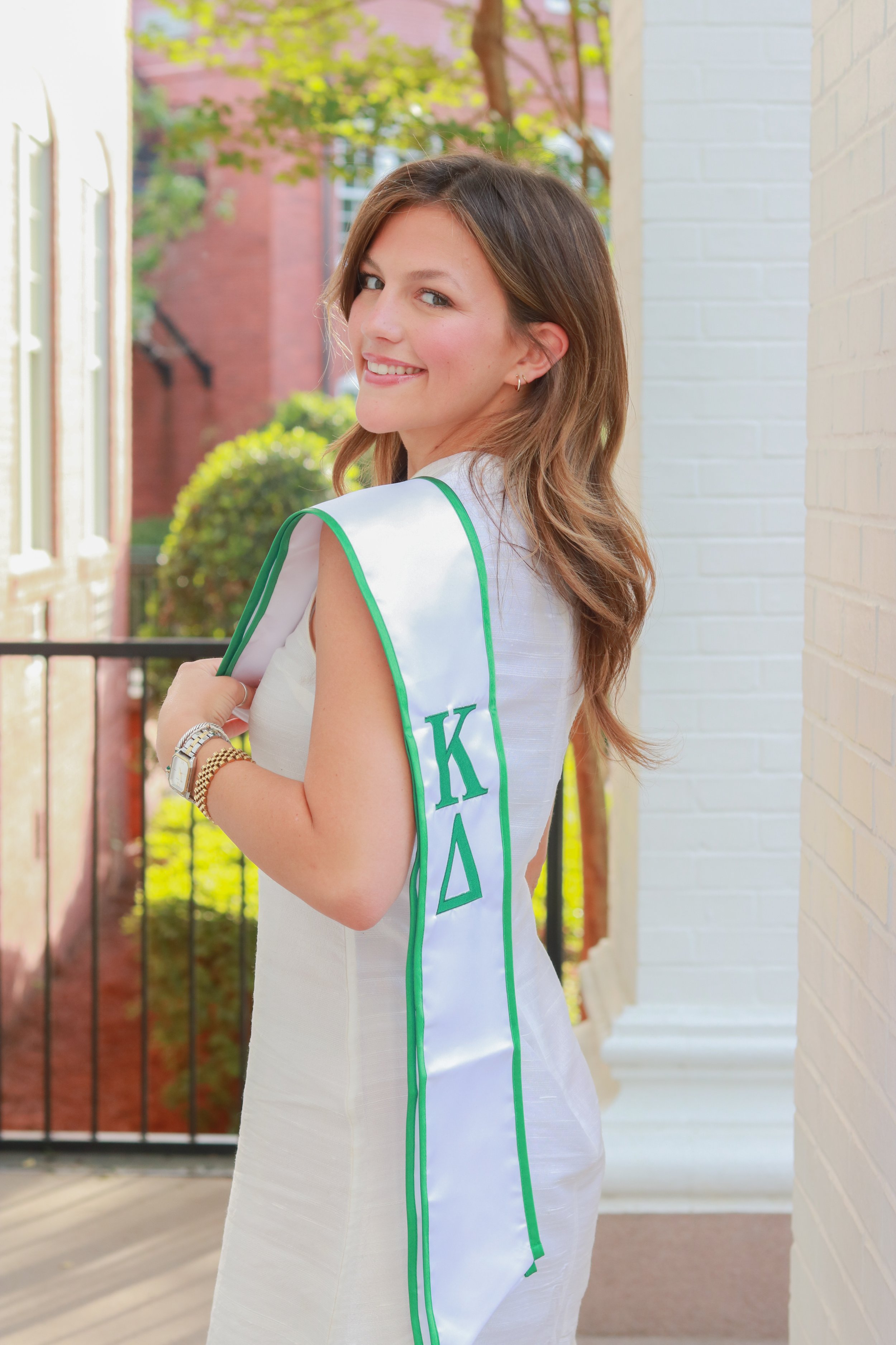 A young woman with long, wavy brown hair and a white dress with green trim, smiling and looking over her shoulder, standing outdoors near a white brick wall with a black iron fence and greenery in the background.