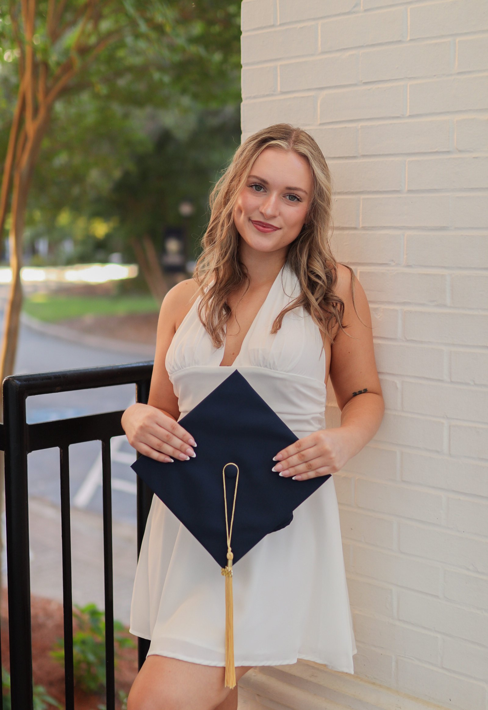 Young woman with wavy blonde hair holding a navy blue graduation cap with a gold tassel, standing outdoors next to a beige brick wall, smiling at the camera.