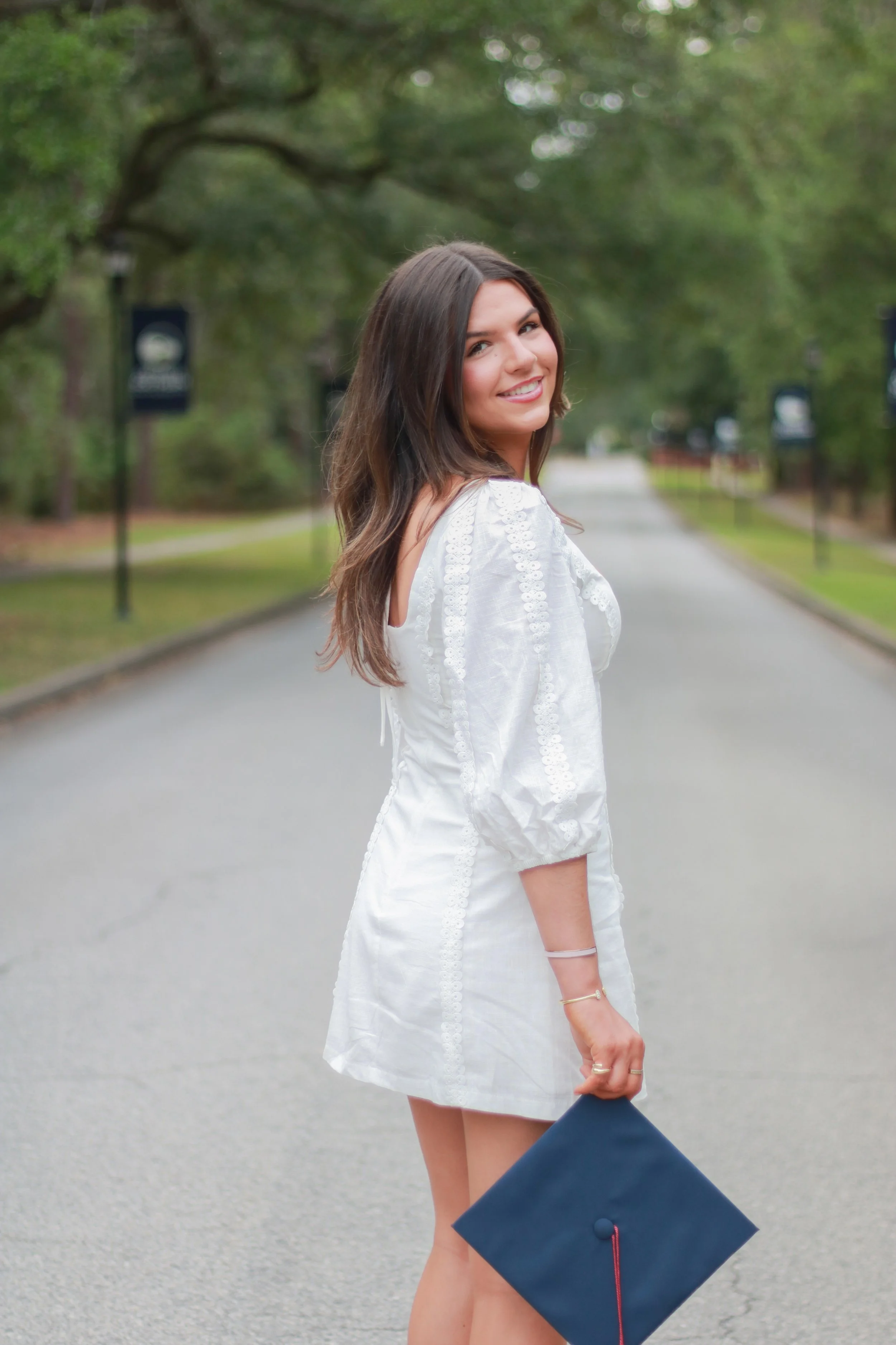 A young woman with long brown hair smiling, dressed in a white embroidered top, standing on a paved pathway lined with trees, holding a navy blue graduation cap.
