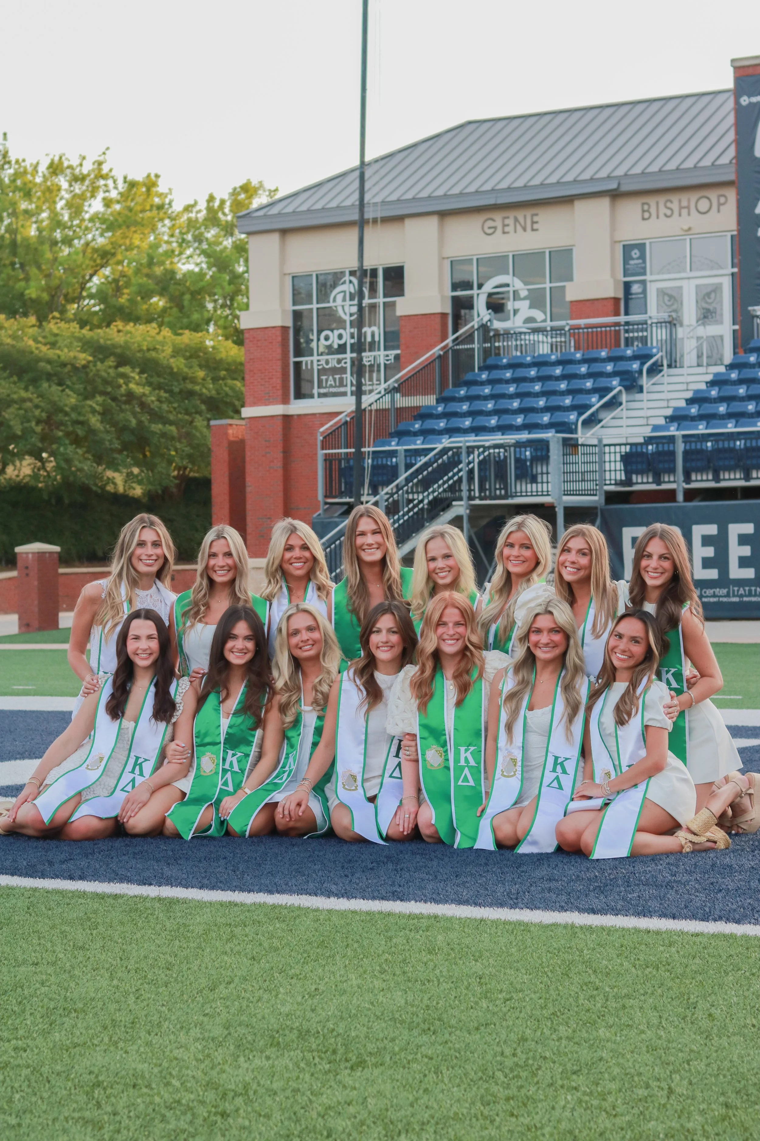 Group of women wearing white and green kappa sorority graduate sashes, posing on a sports field with stadium seating and a building in the background.