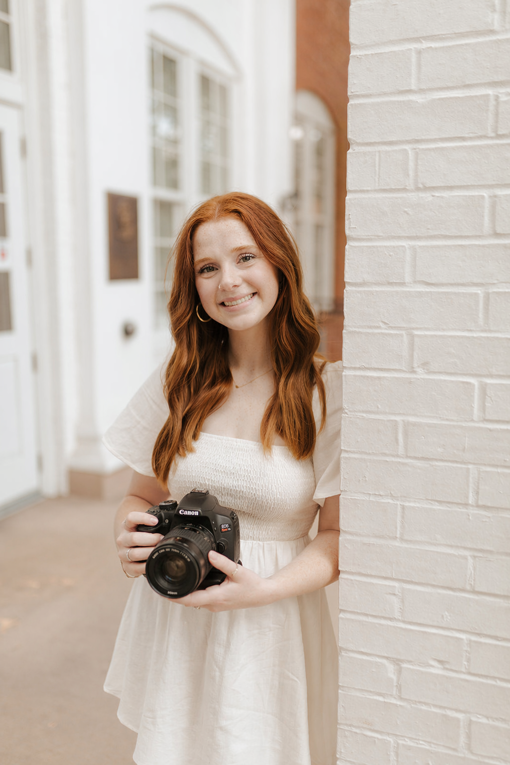 A young woman with red hair and a white dress smiling while holding a camera, standing next to a white brick wall in an outdoor urban setting.