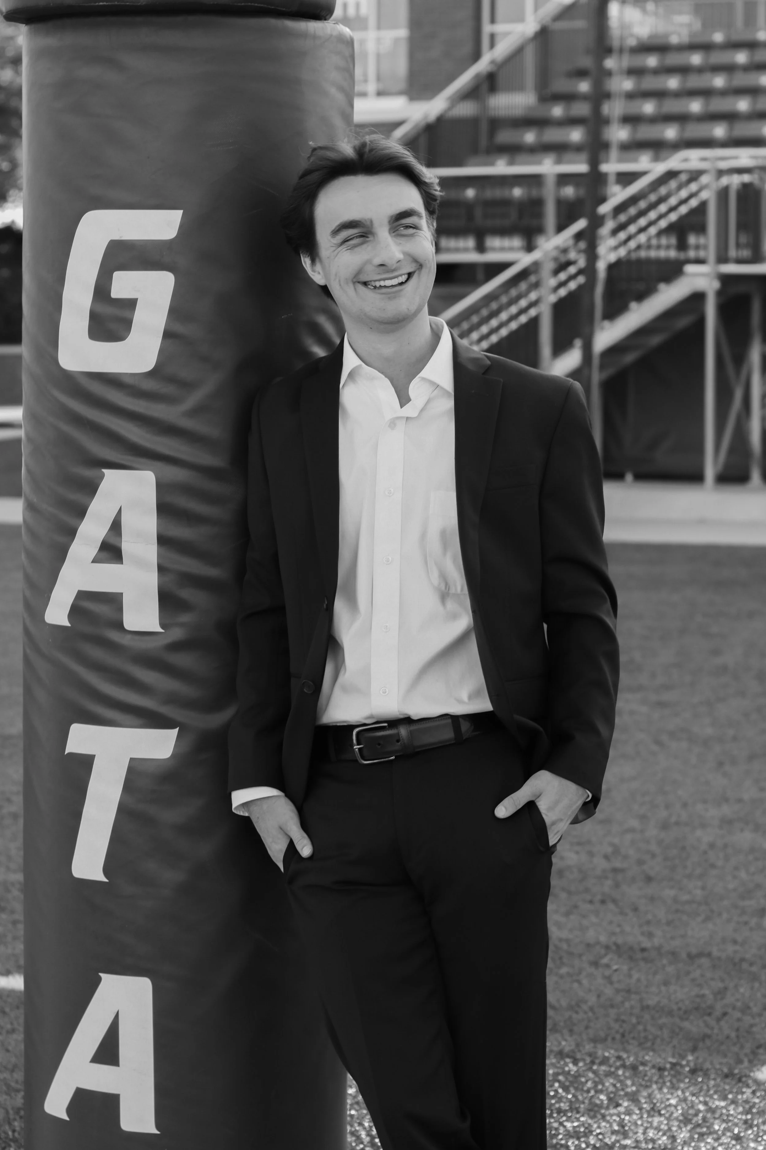 A young man in a suit and white shirt smiling and leaning against a large vertical pole marked with the letter 'A' in a city or outdoor setting.