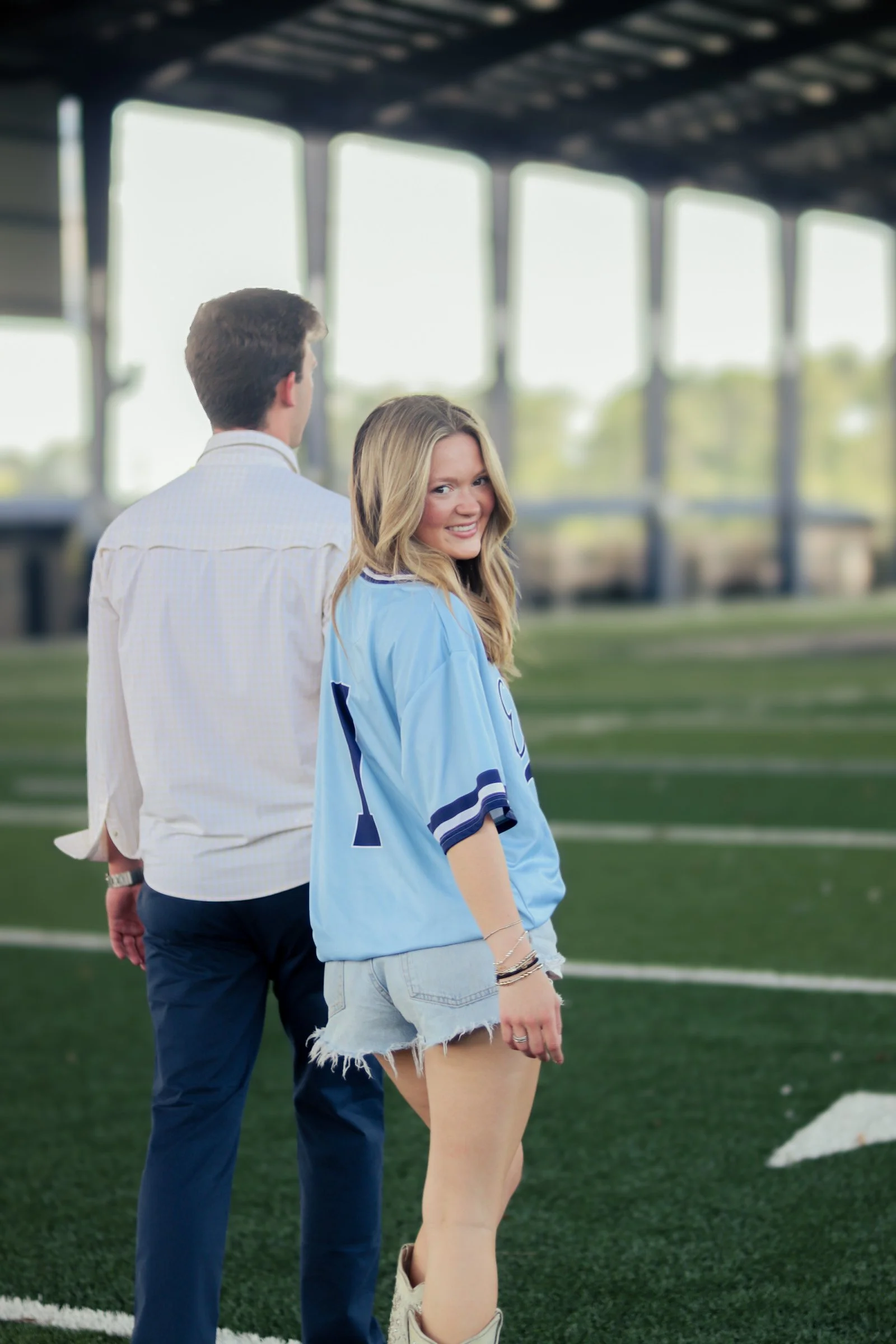 A young woman smiling, wearing a light blue sports jersey with the number 2 on it, standing on a green football field with a man in a white shirt and blue pants behind her. They are near the sideline with large windows and stadium seating in the back