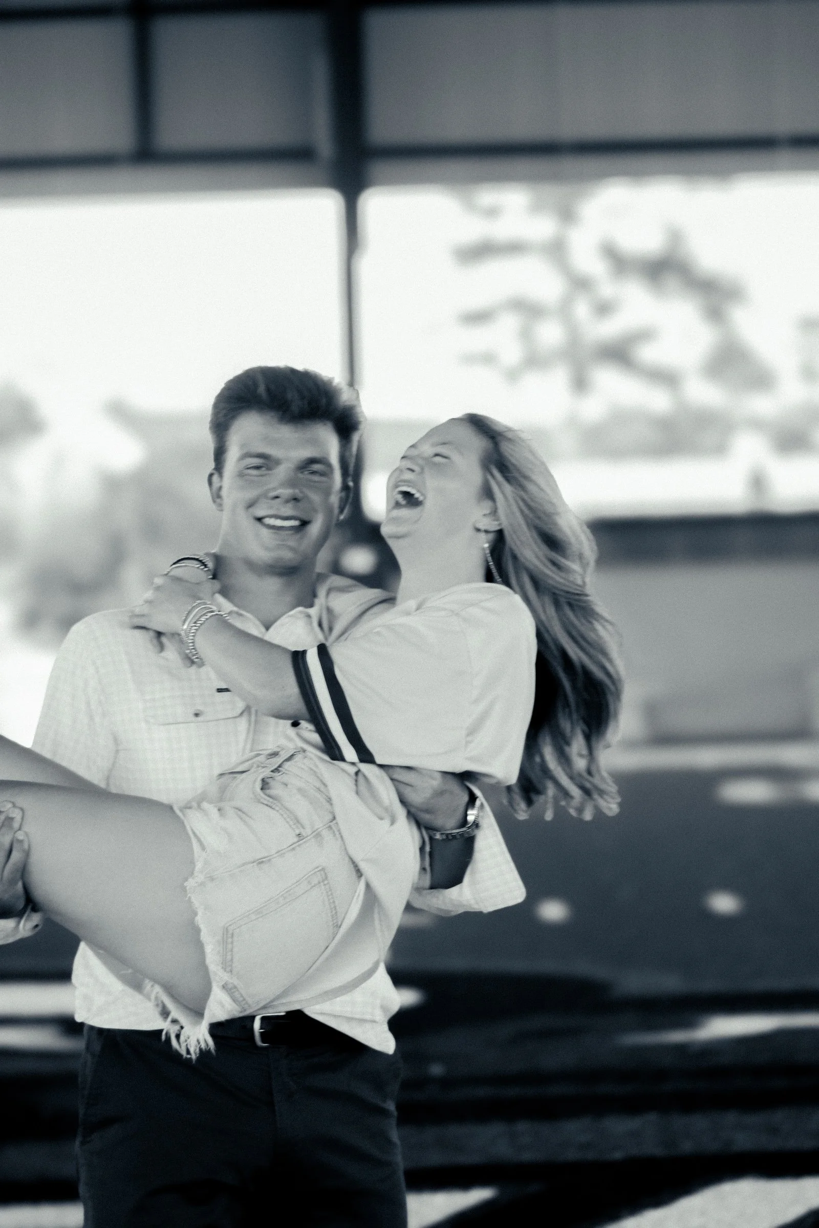 A man carrying a woman in his arms, both smiling and laughing, at an outdoor location with trees and cars in the background.