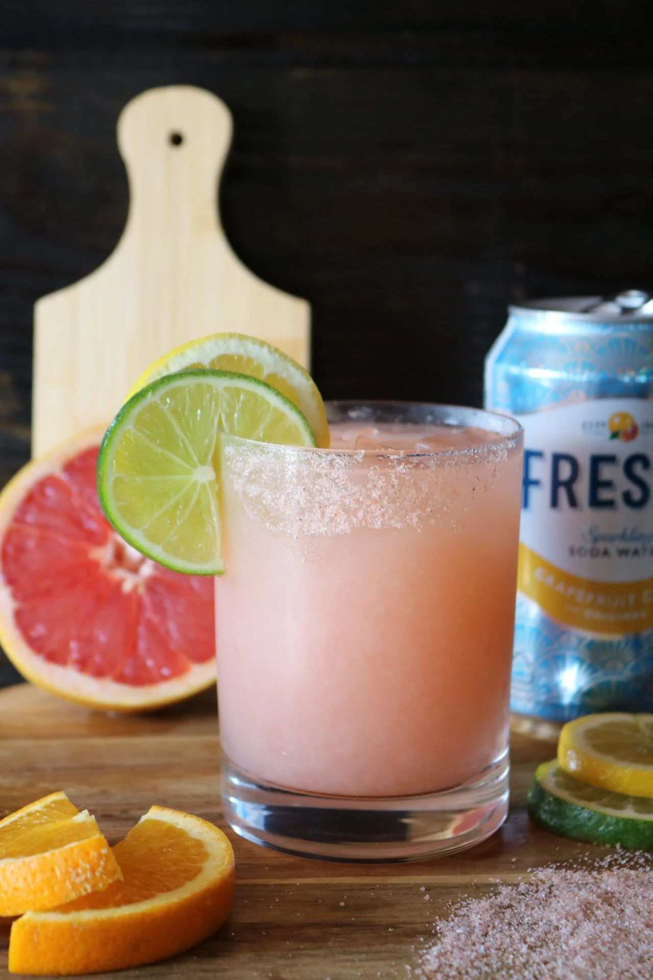 A pink grapefruit cocktail garnished with slices of lemon and lime. The drink is in a clear glass with salt around the rim. Behind the drink, there are halves of grapefruit and slices of orange, lemon, lime, and cucumber, along with a can of sparklin