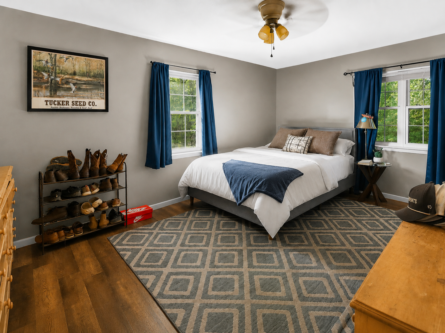 A bedroom with a bed, two windows with blue curtains, a ceiling fan, a patterned rug, a shoe rack, and a wooden dresser.