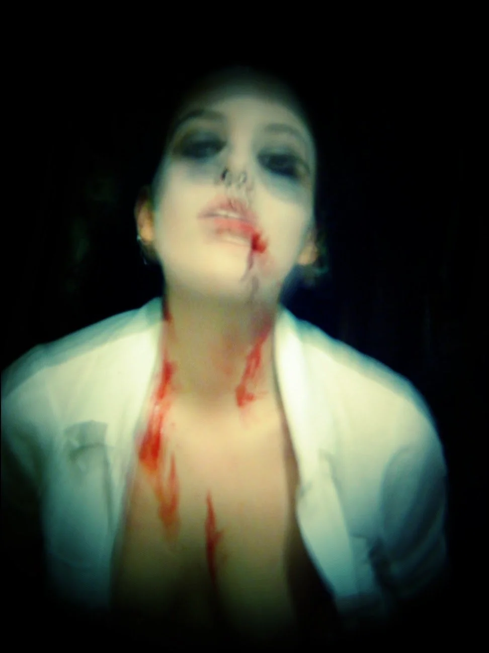 The image features a female vampire, with dark eye makeup and black lipstick. Blood appears to be dripping down from her mouth and neck, and her shirt is open at the collar. The background is black, giving a haunting or horror-themed appearance.