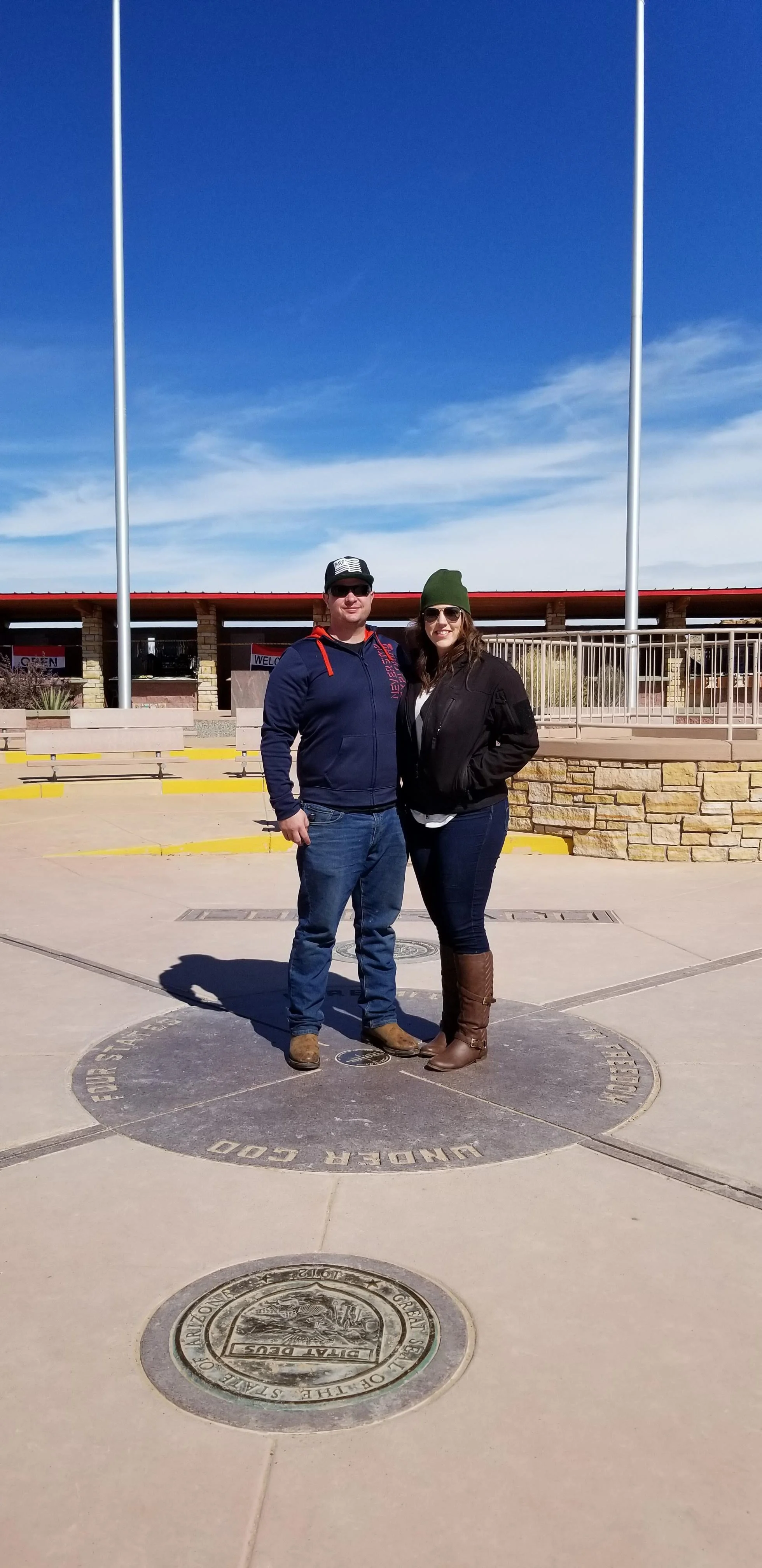 Two people standing on a geographic marker at the border of Utah and Colorado, with a clear blue sky in the background.