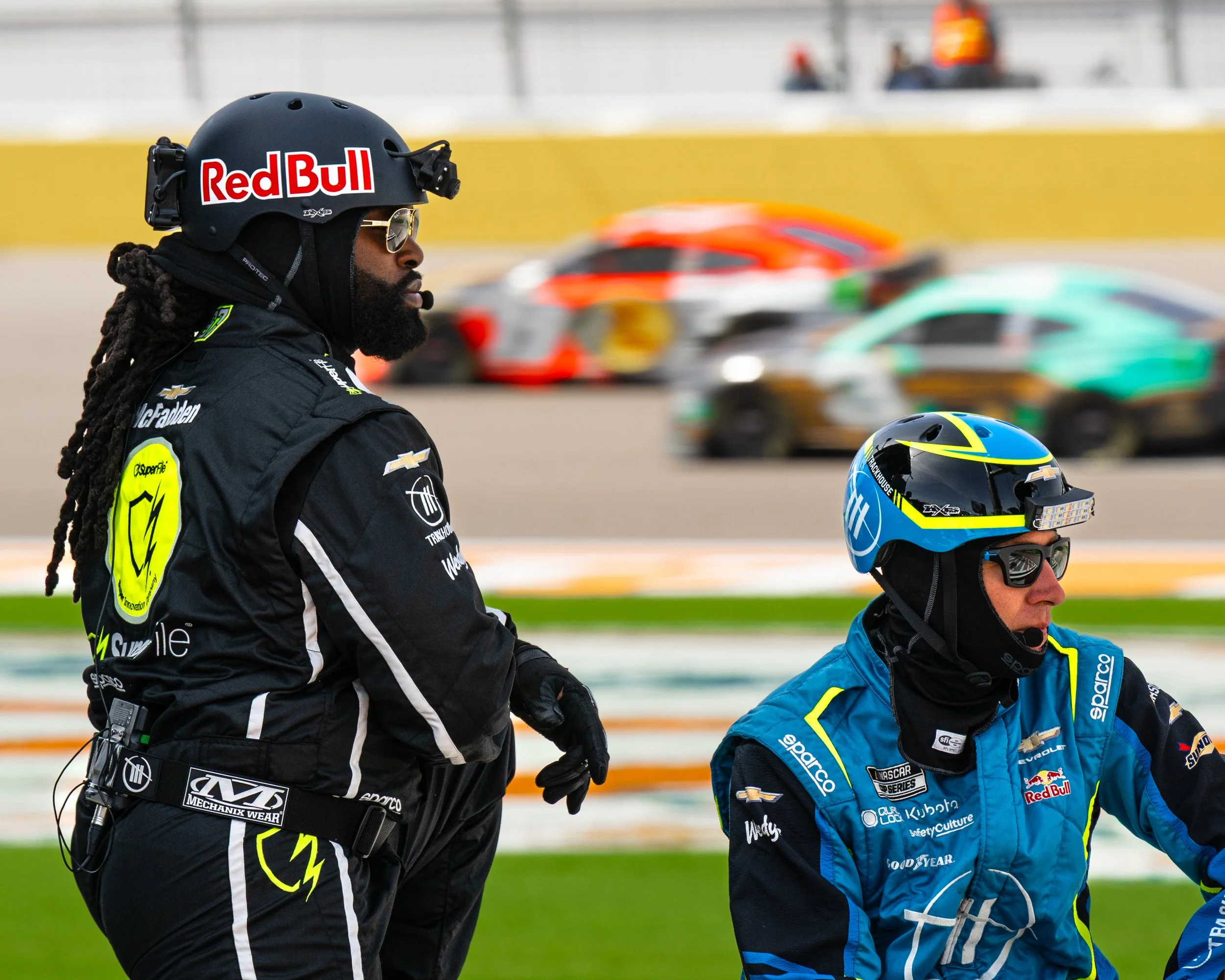 Two race car drivers wearing racing suits and helmets walking on the NASCAR Las Vegas race track with a blurred background of race cars and spectators.