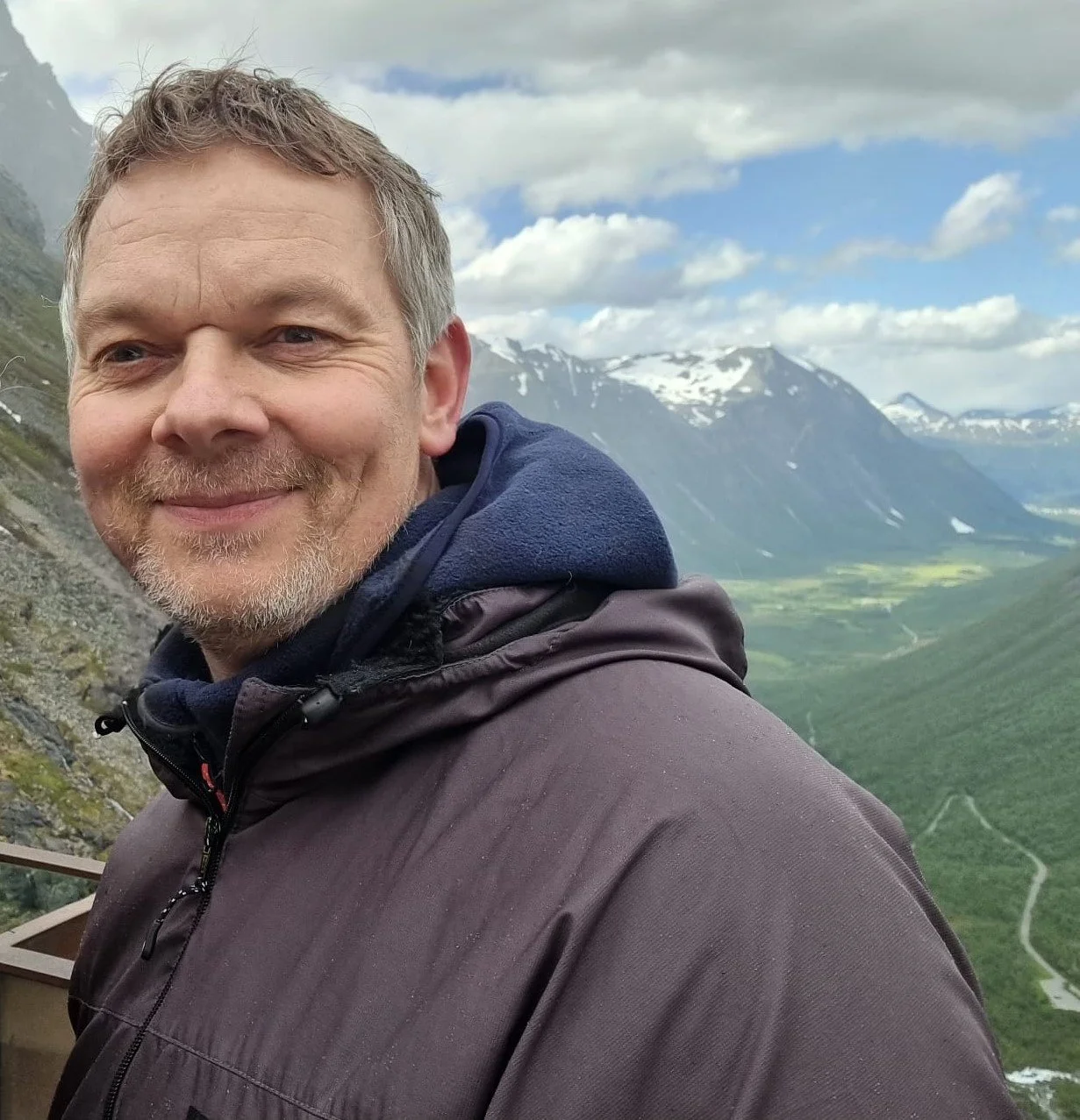 A man smiling outdoors with a mountain landscape, snow-capped peaks, and a valley with winding roads in the background.