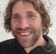 Close-up of a man with wavy brown hair and a beard, smiling.