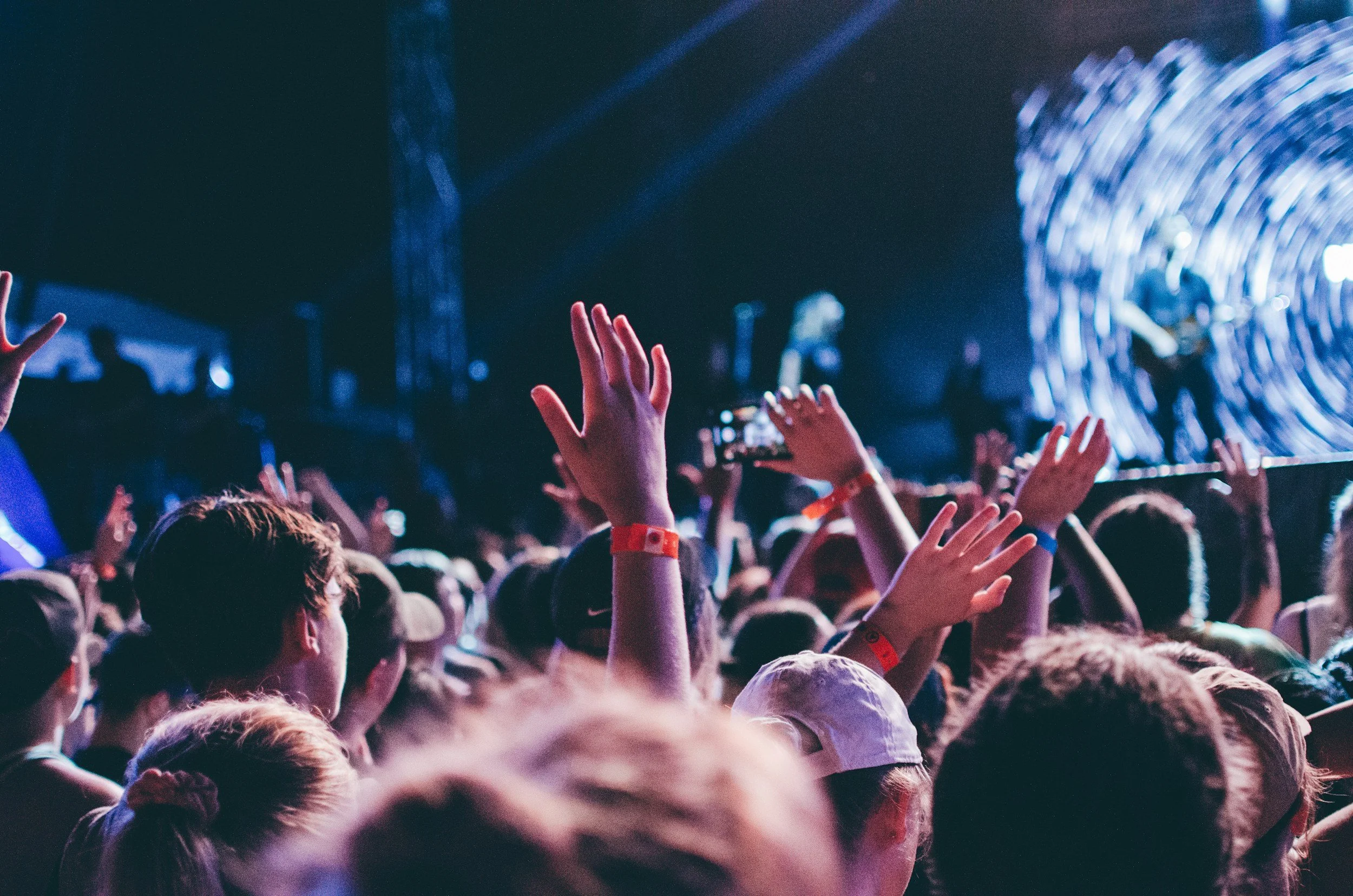 Crowd of people with raised hands at a concert or festival with stage lights and a large digital screen in the background.