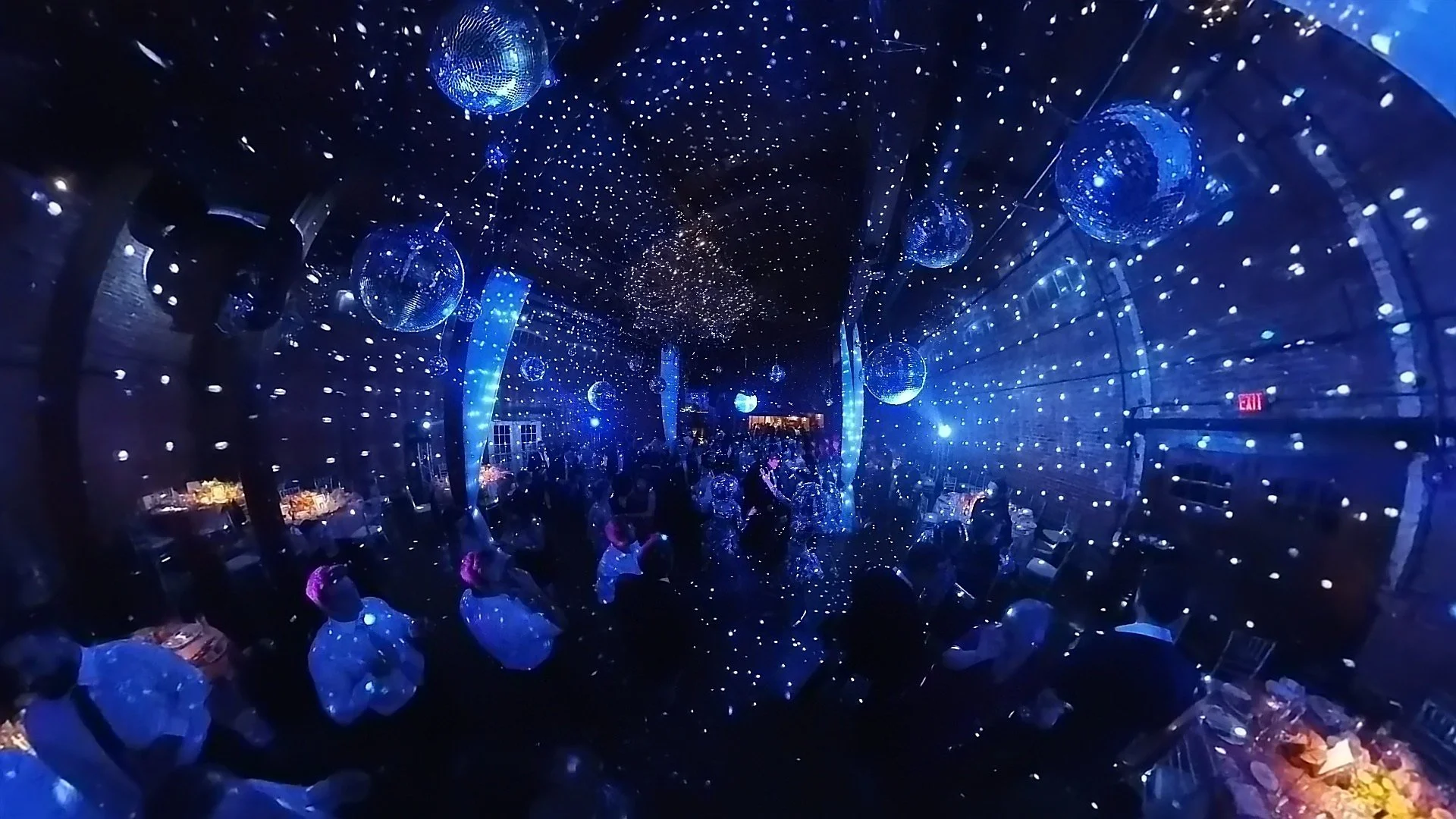 A dimly lit elegant event space illuminated by numerous small blue LED lights on the ceiling, with several large disco balls hanging and reflecting light across the room. The room is filled with people sitting at tables, engaging in conversations, with some standing and moving around.