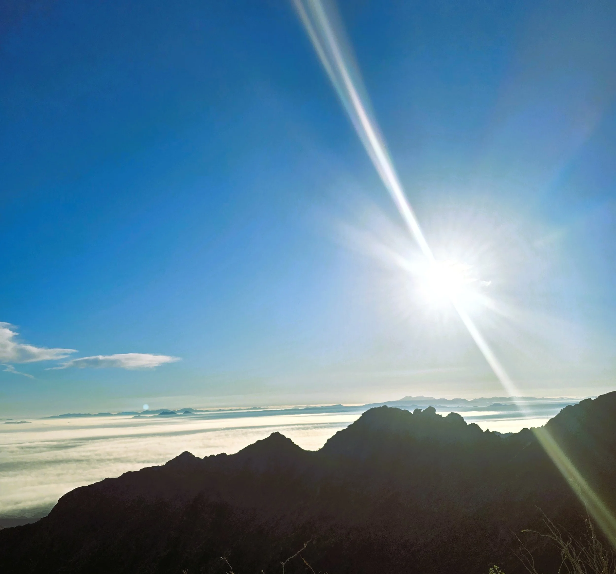Scenic view of a coastal landscape with mountains and the ocean, taken during sunrise or sunset, with a bright sun flare at the bottom right.