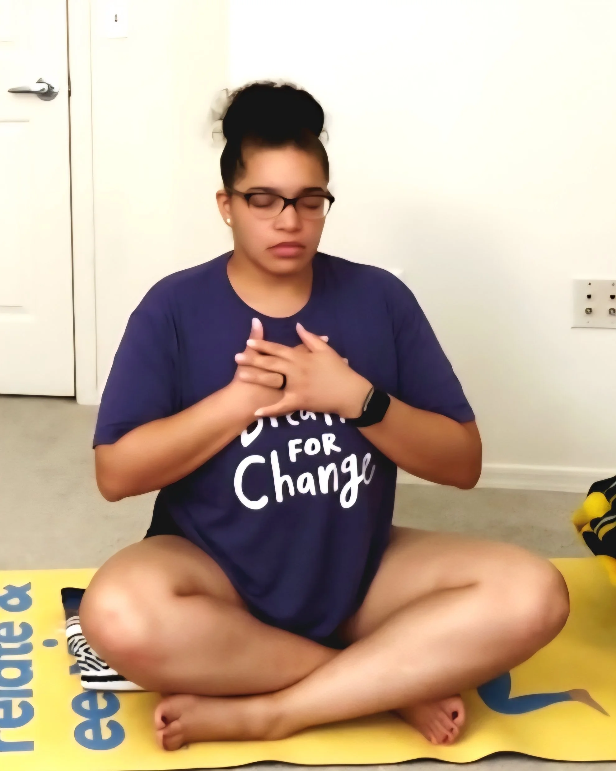 A young woman with glasses and earrings, wearing a blue shirt, sitting cross-legged with hands on her chest in front of a white wall and electrical outlet, with part of a yellow educational sign visible. She has her eyes closed.