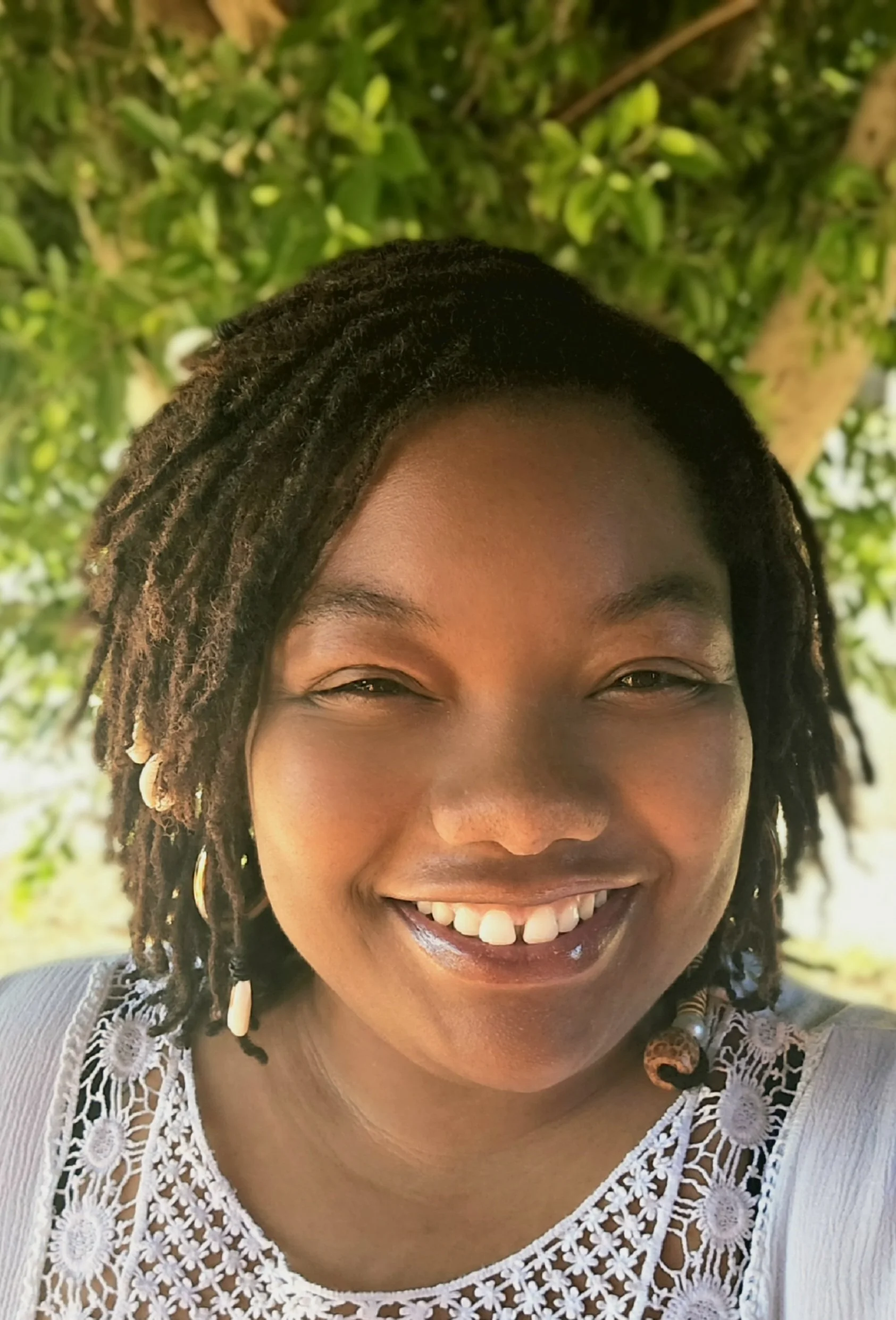 A smiling woman with dark hair styled in dreadlocks, wearing a white lace top, standing outdoors with green foliage in the background.