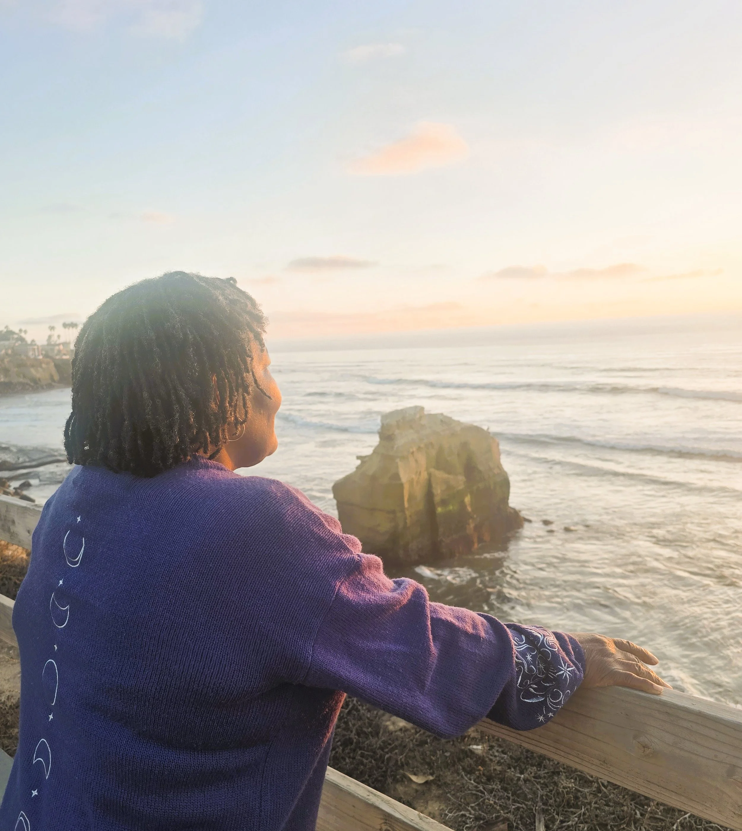 A person with curly hair wearing a purple hoodie sitting on a wooden railing while looking at the ocean during sunset.