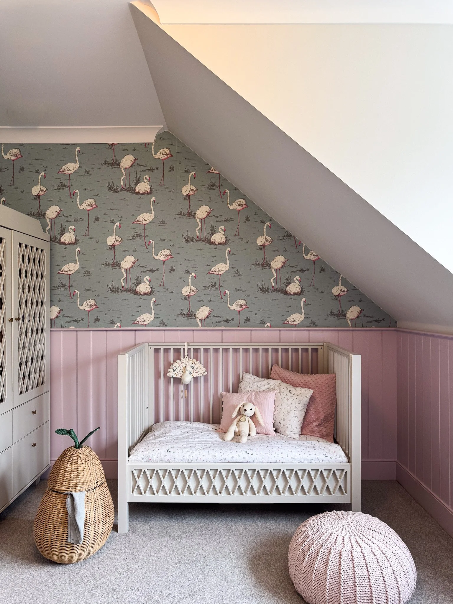 Designing a child's room well means thinking ten years ahead. The Cole and Son Flamingos wallpaper in sage is installed across the pitched ceiling and upper walls. Pink tongue-and-groove grounds it below. The lattice wardrobe provides storage that wi