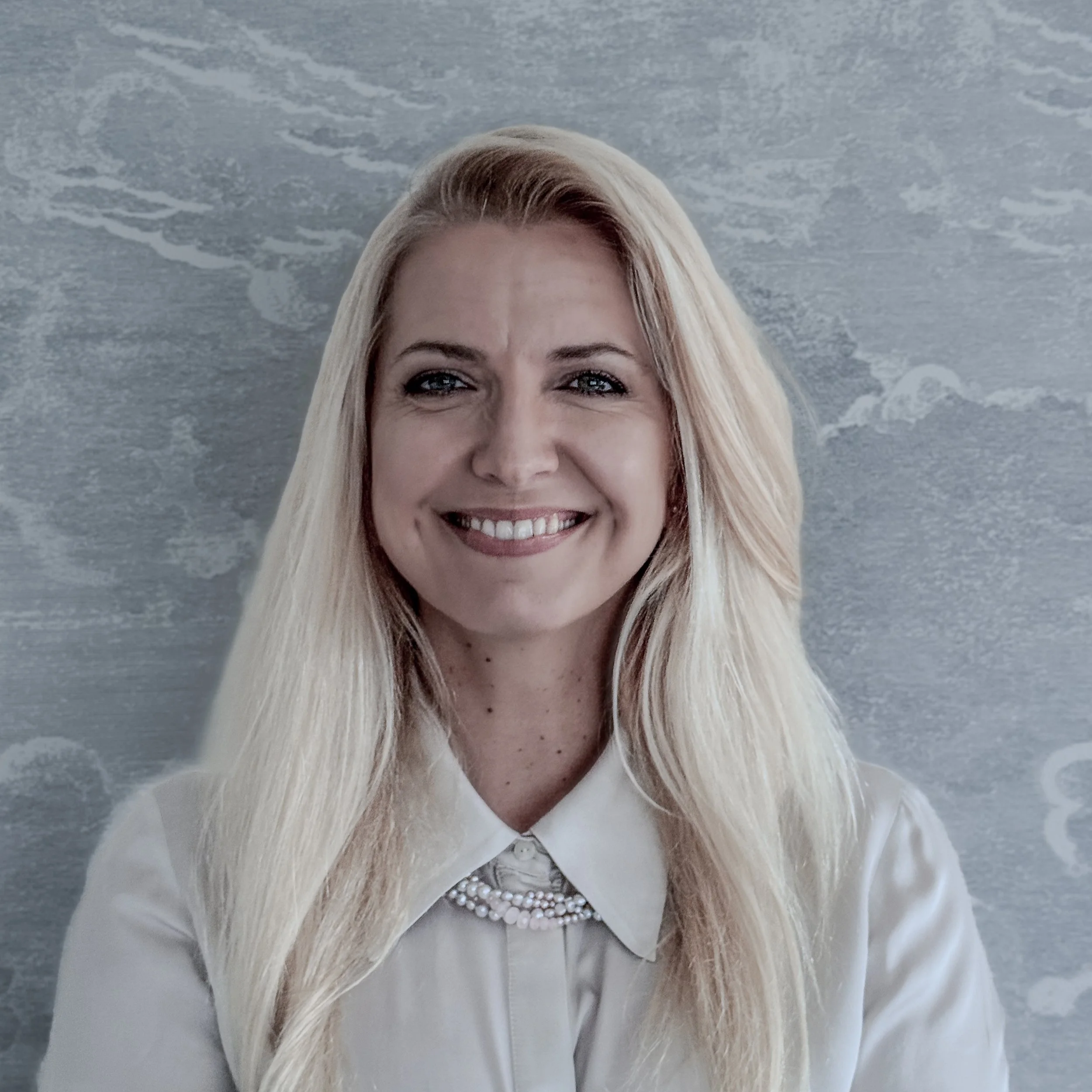 Close-up of a smiling woman with blonde hair, wearing pearl jewelry and a white outfit, standing in front of a textured grey background.