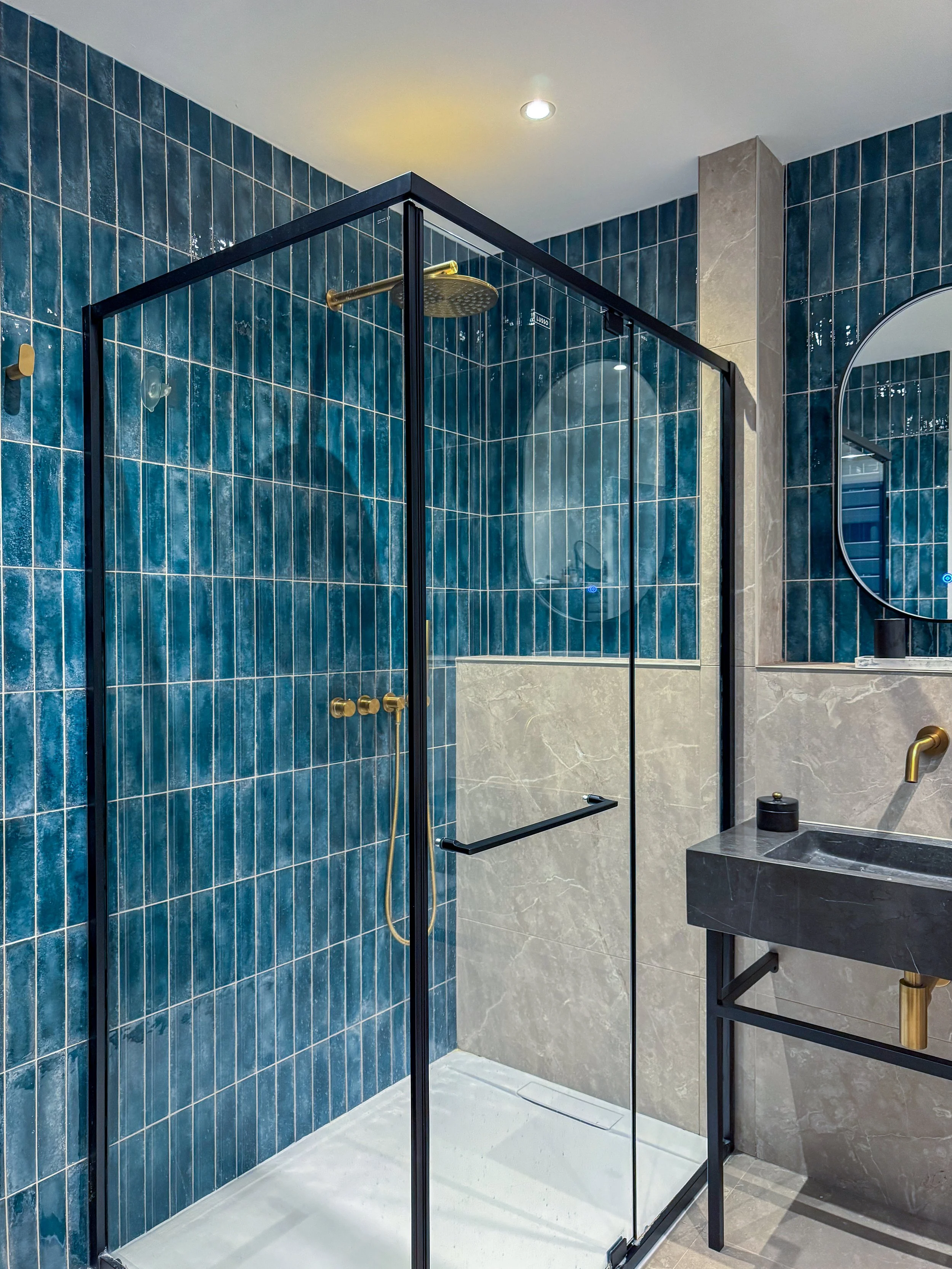 The same deep teal zellige carries through from the cloakroom, a deliberate thread connecting two spaces. Here it is paired with warm grey stone-effect tile, a black steel shower frame, brushed brass fittings and a honed dark marble basin on an open 