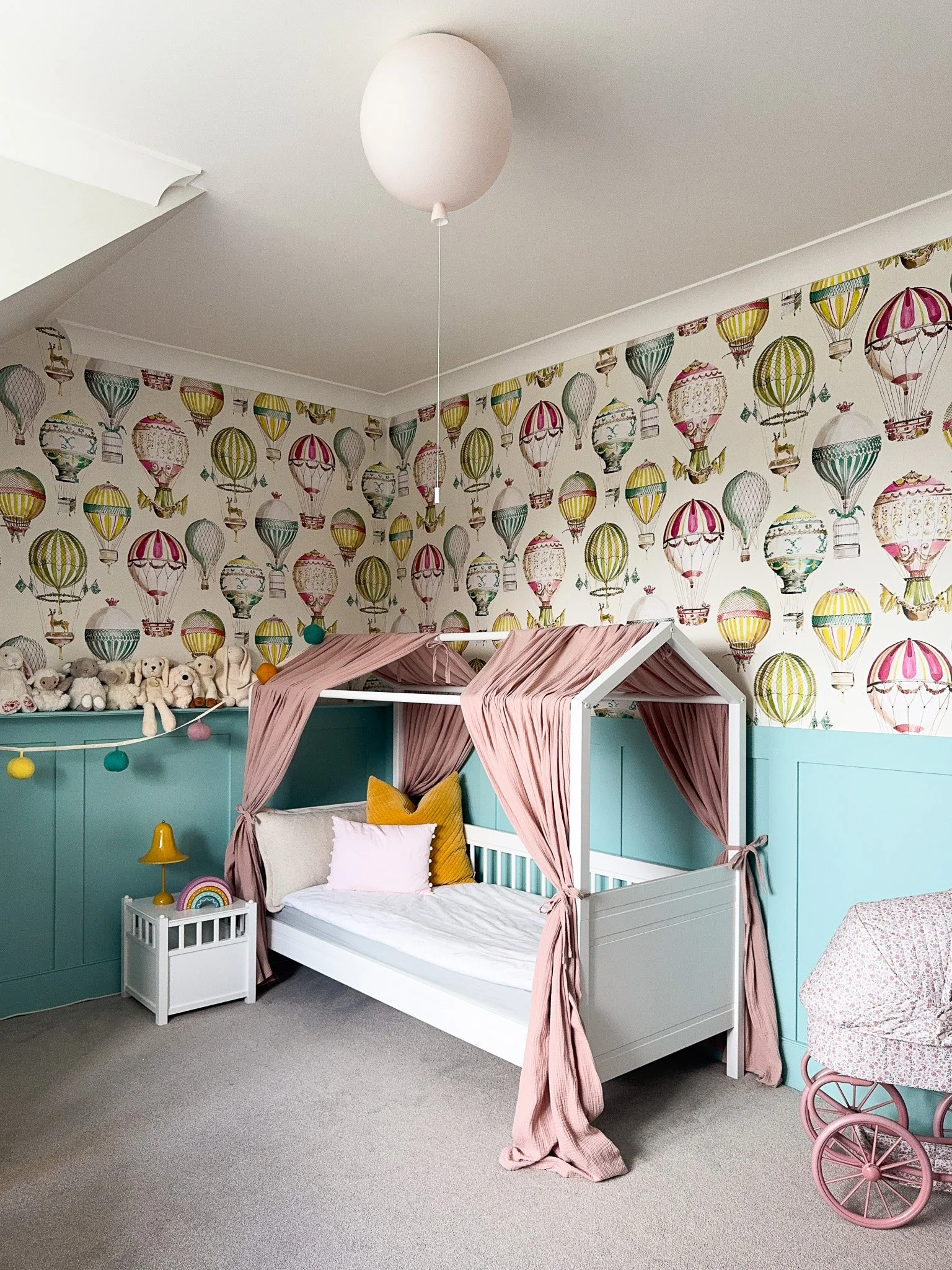 A house-frame bed with dusty pink linen canopy. Hot air balloon wallpaper in full colour. Teal panelling that anchors the pattern without competing with it. A balloon pendant that completes the story without being cartoonish. This room works because 